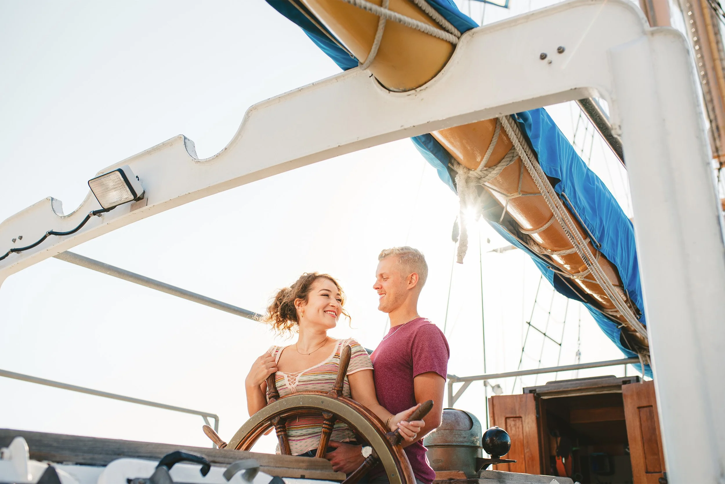 Couple smiling at each other at the helm of a sailboat, nautical engagement photoshoot Orange County