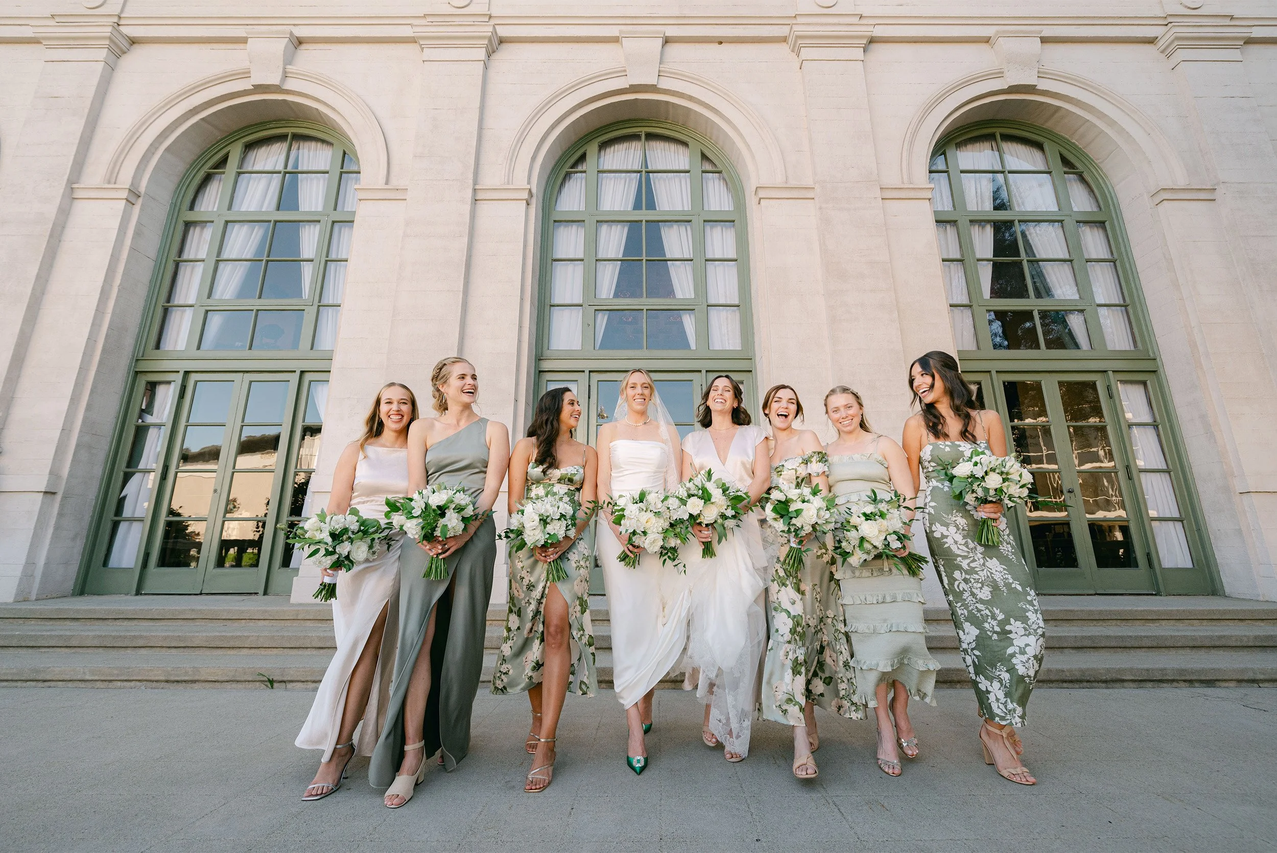 Award-winning wedding photographer in Orange County captures bride and bridesmaids laughing together in sage green and floral dresses outside The Ebell of Los Angeles