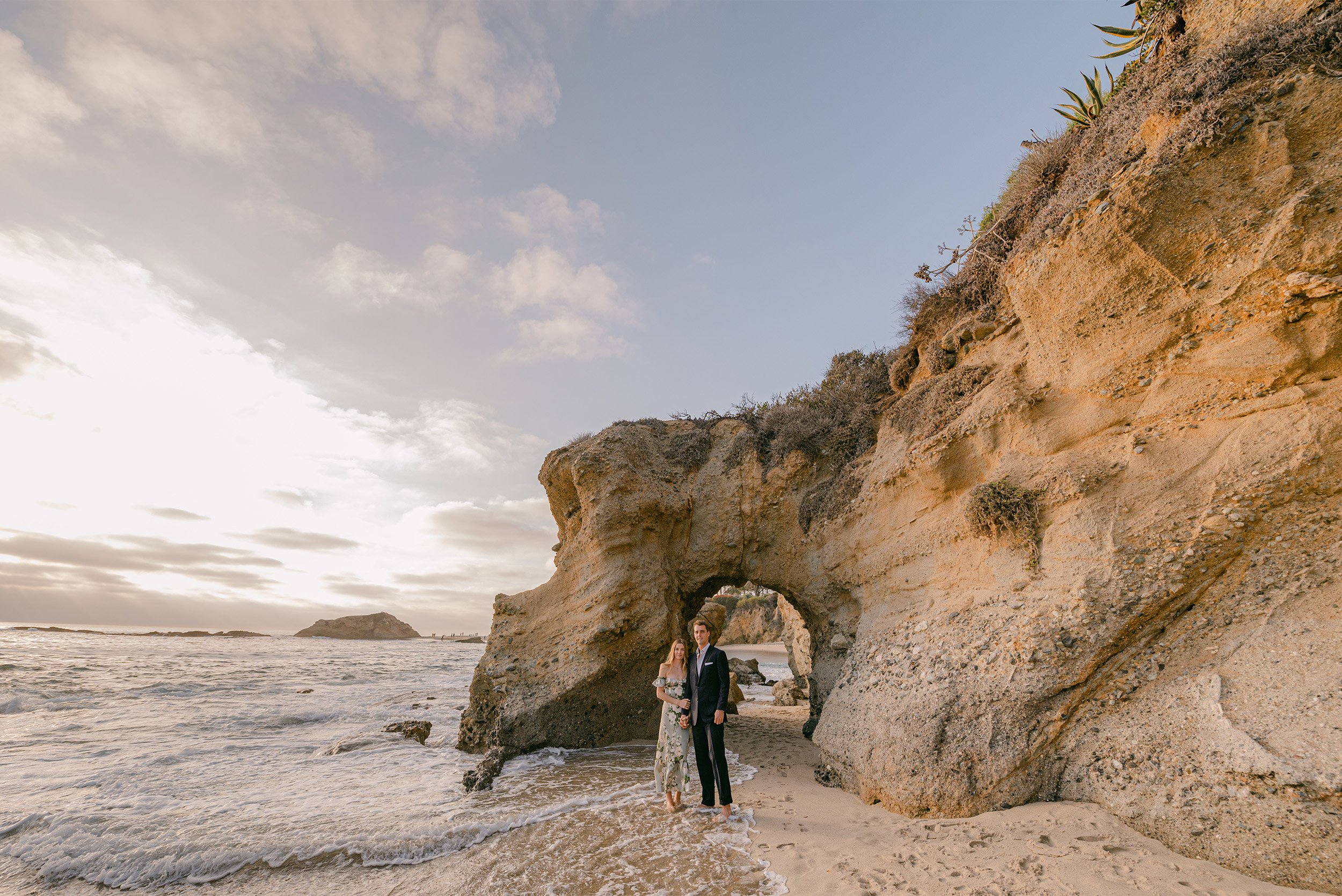 Couple standing under natural rock arch at Laguna Beach during sunset, award-winning engagement photographer