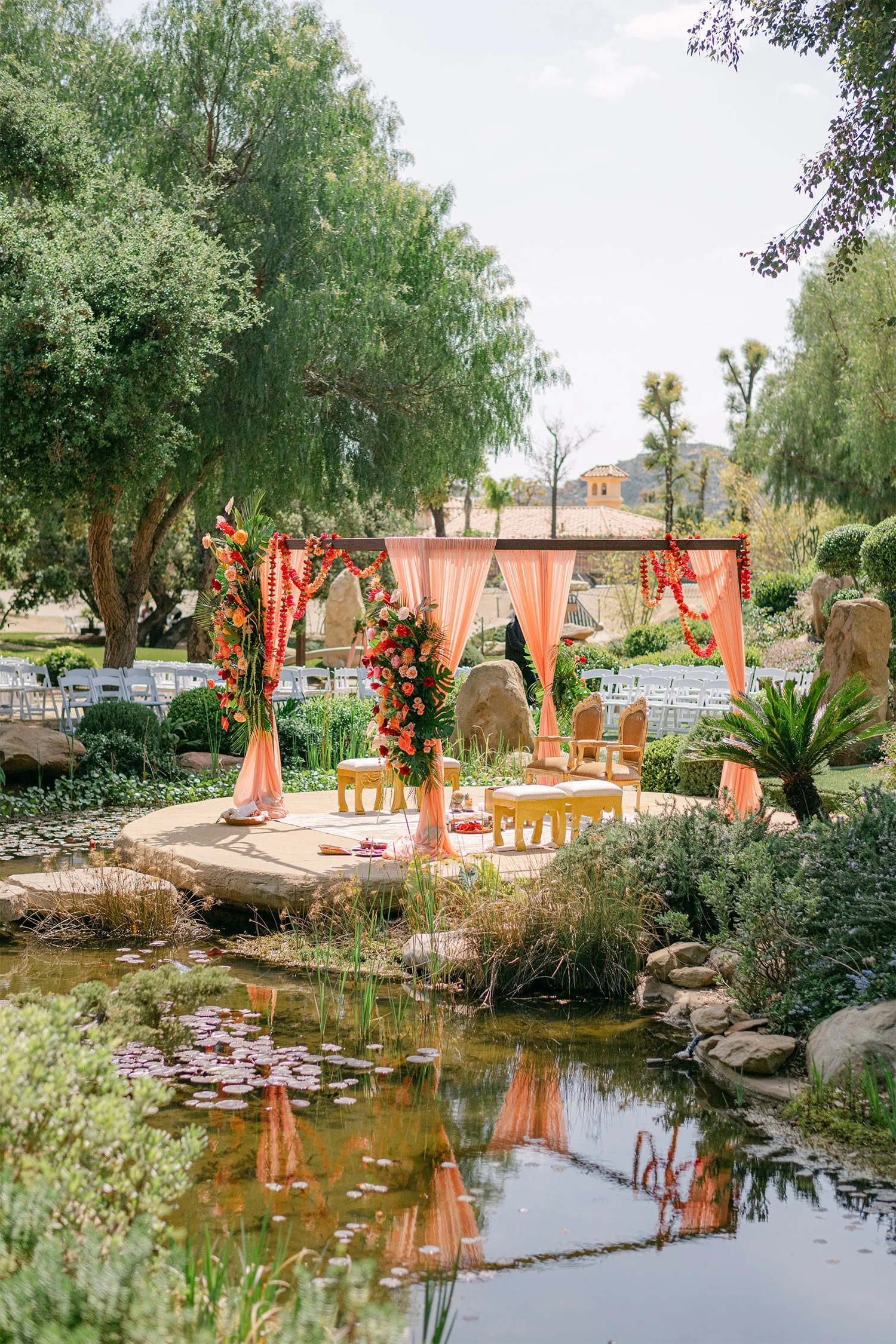 Outdoor wedding ceremony setup with peach draping and floral arch beside koi pond, Indian wedding decor