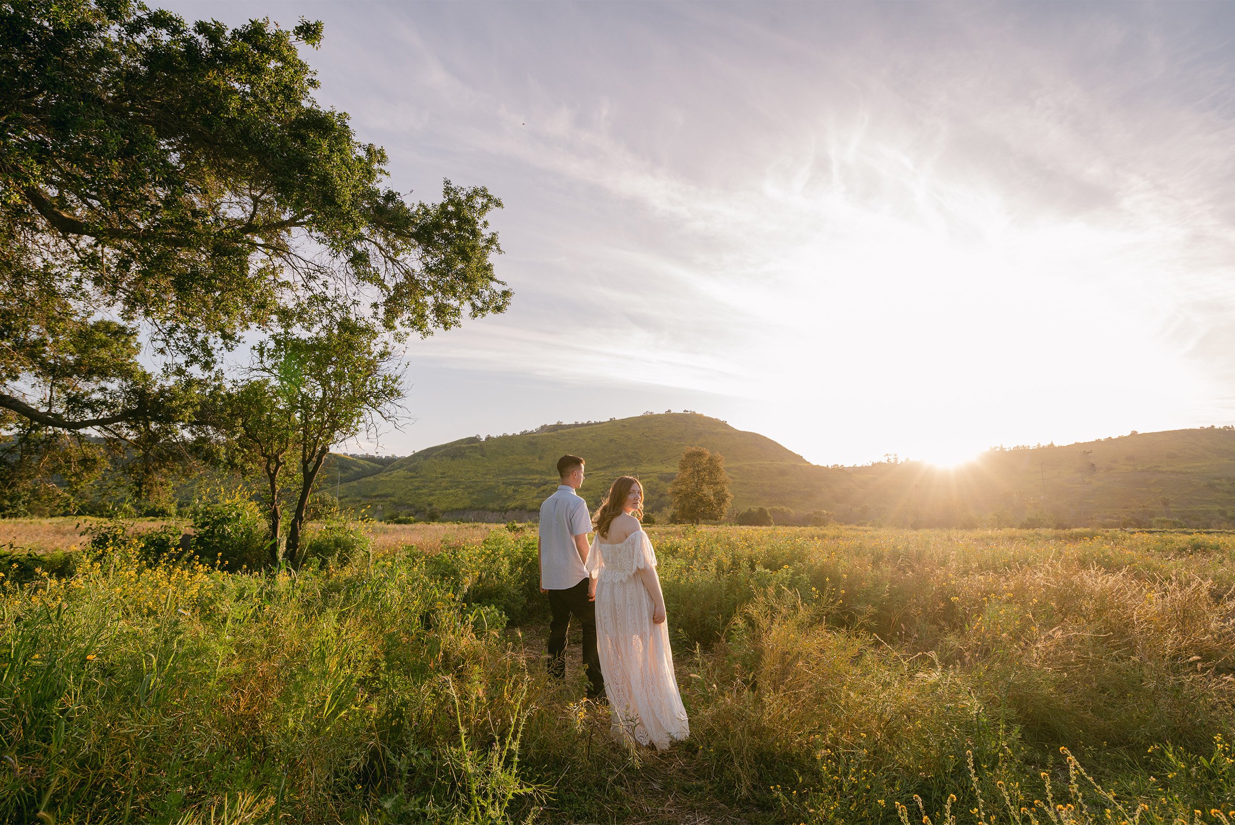 Pregnant couple walking through golden wildflower meadow at sunset, award-winning maternity photographer in Orange County