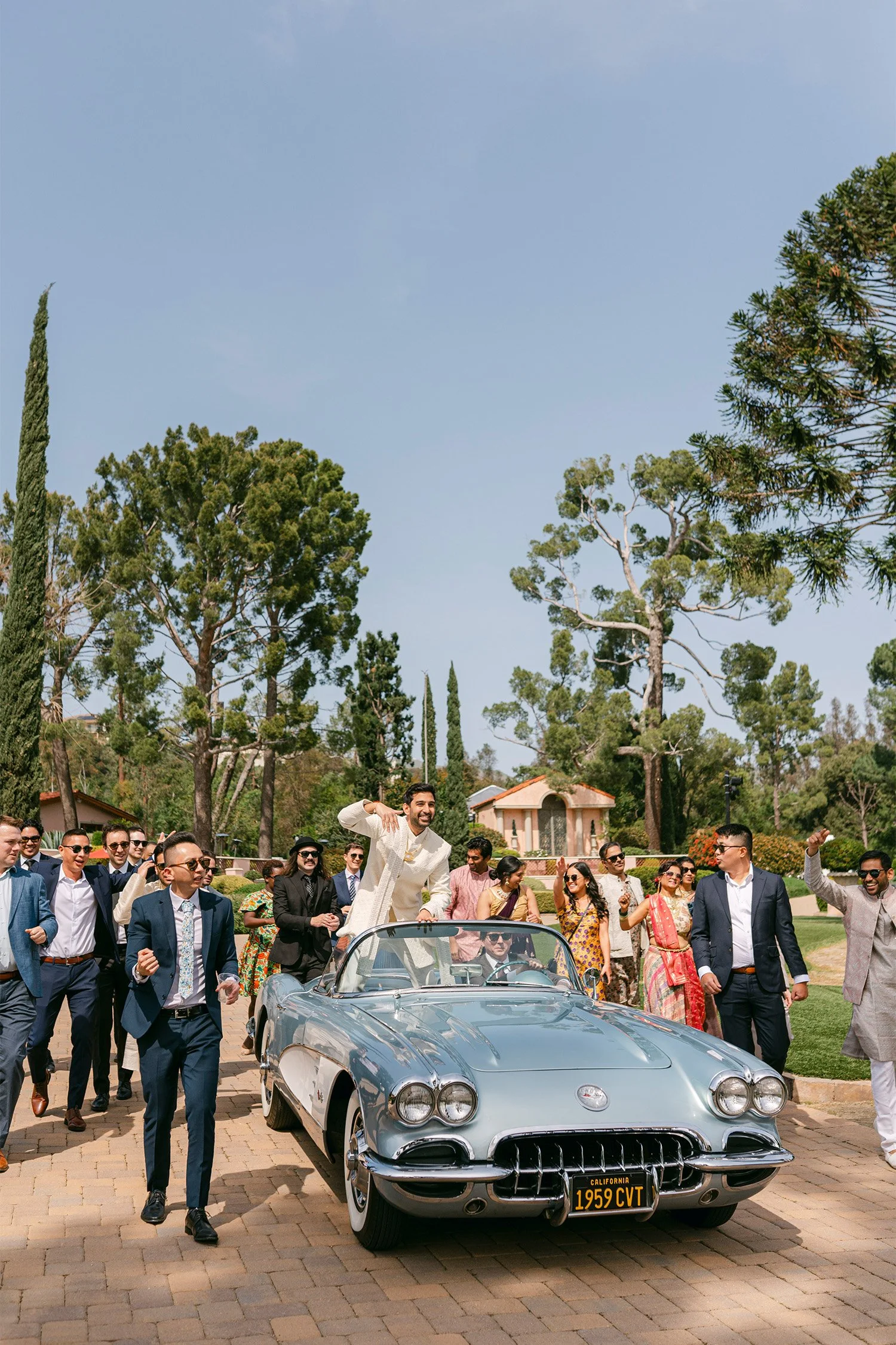 Groom celebrating on vintage Corvette convertible surrounded by dancing wedding guests, Indian wedding baraat