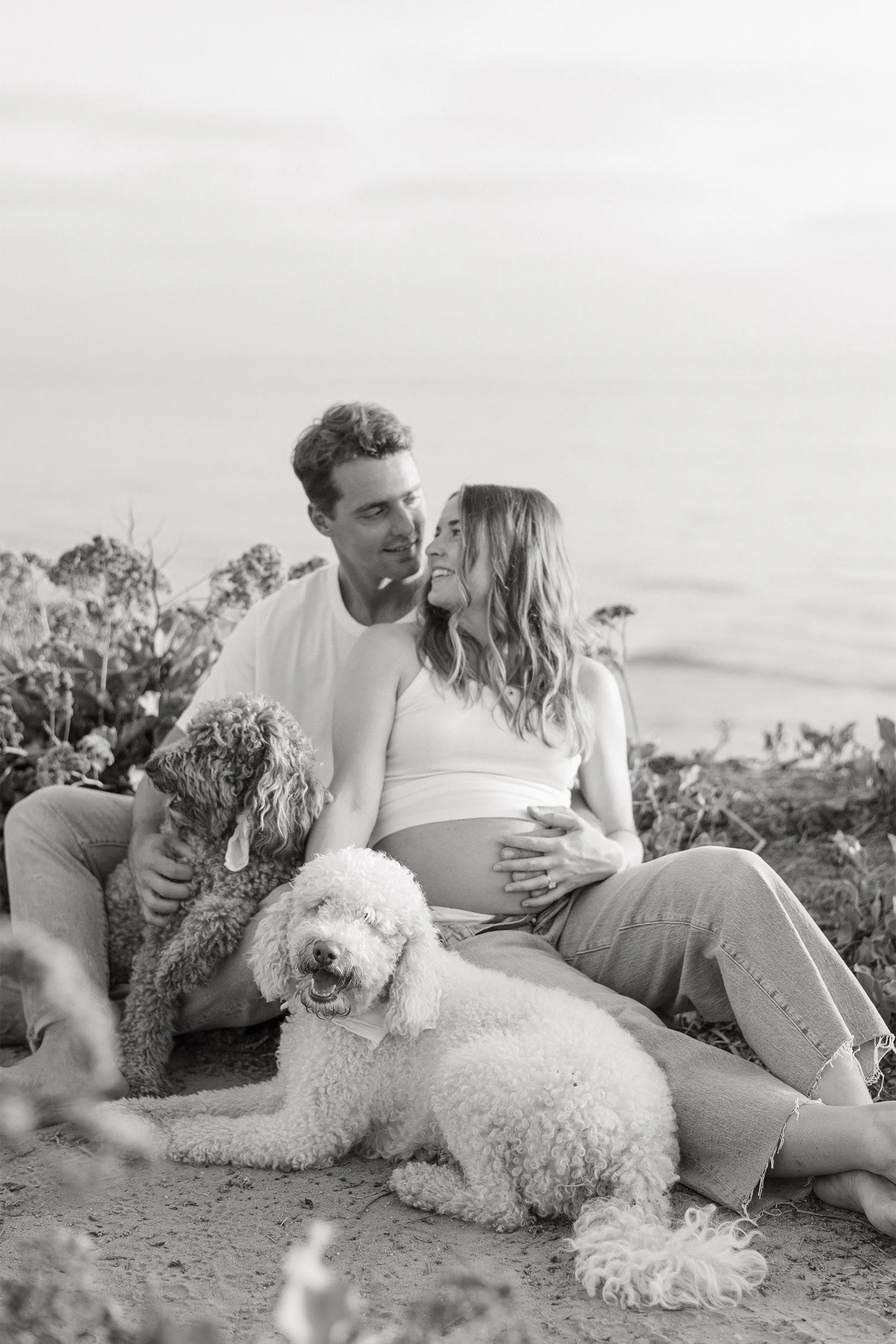 Black and white photo of pregnant couple with two dogs on beach, Orange County pet-friendly maternity photographer