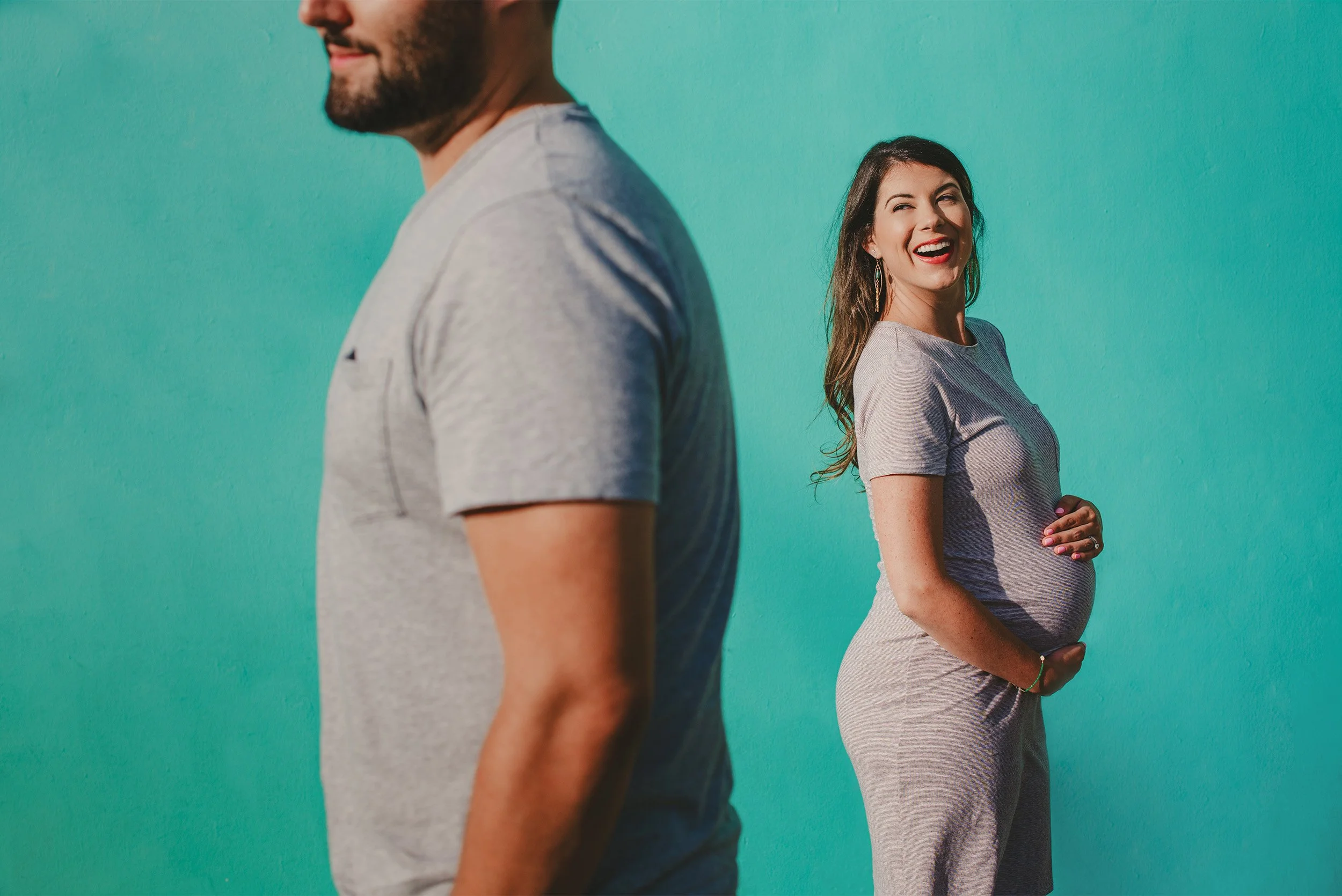 Orange County maternity photographer captures pregnant woman laughing beside husband against vibrant teal wall