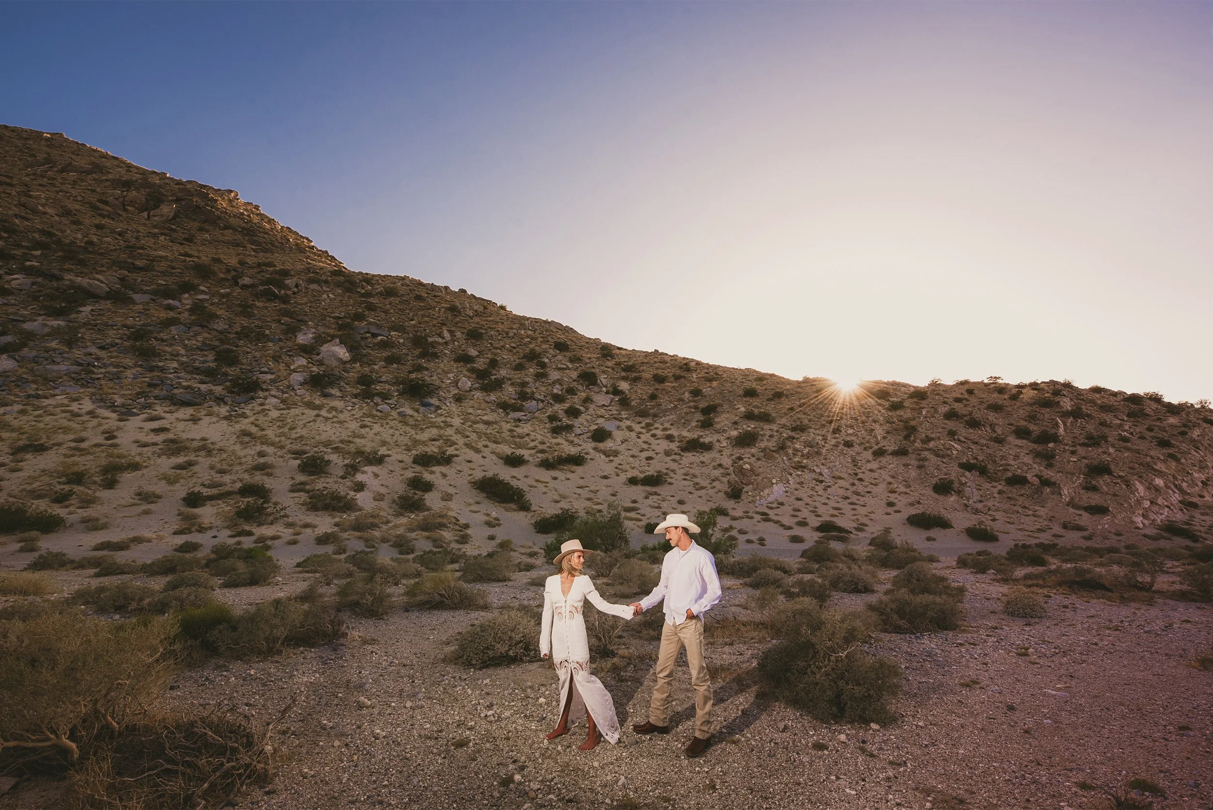 Couple in white western attire holding hands walking through the desert at sunset, Palm Springs cowboy-style engagement shoot