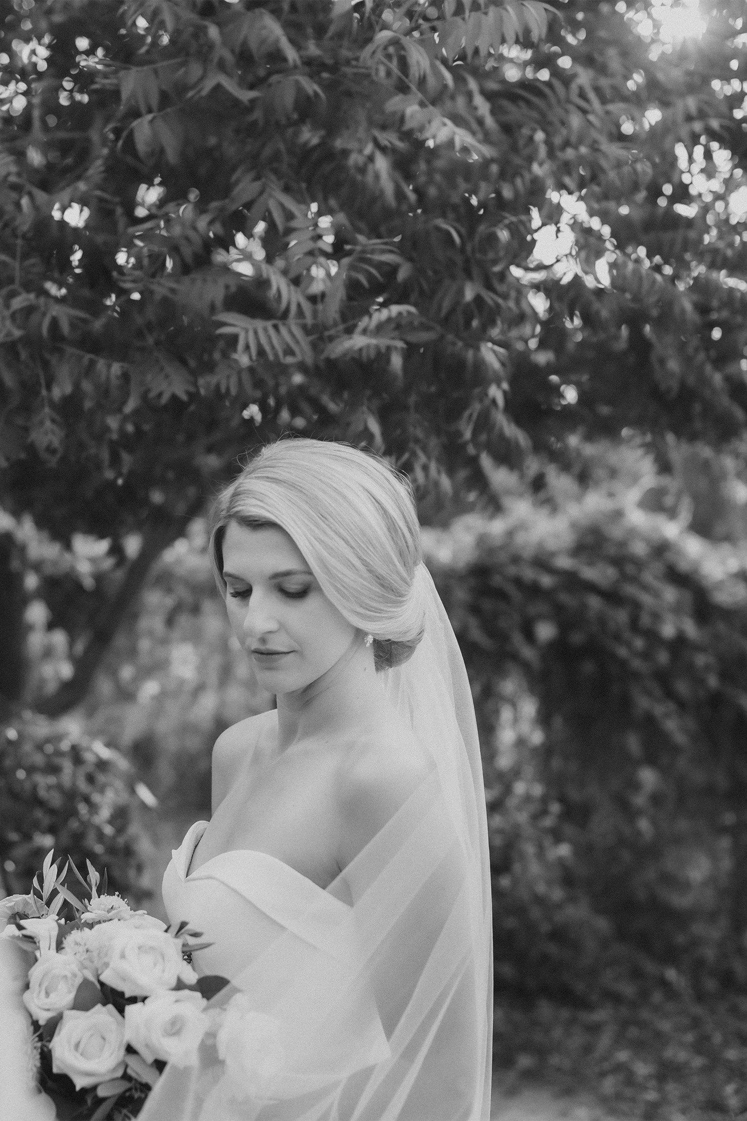 Bride in strapless gown looking down at bouquet under tree canopy, black and white bridal portrait