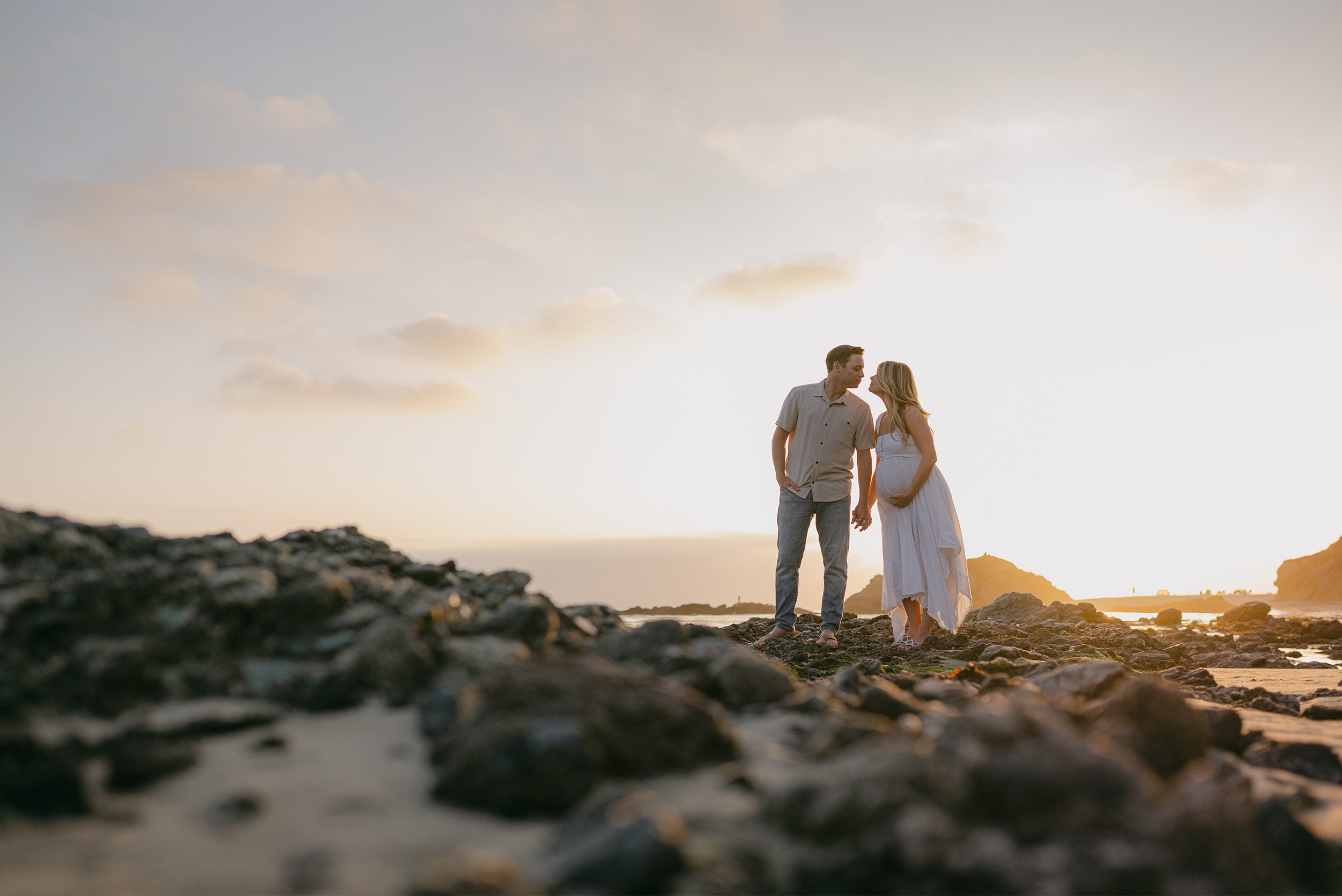 Couple standing close on rocky beach at golden hour, highly recommended maternity photographer Orange County