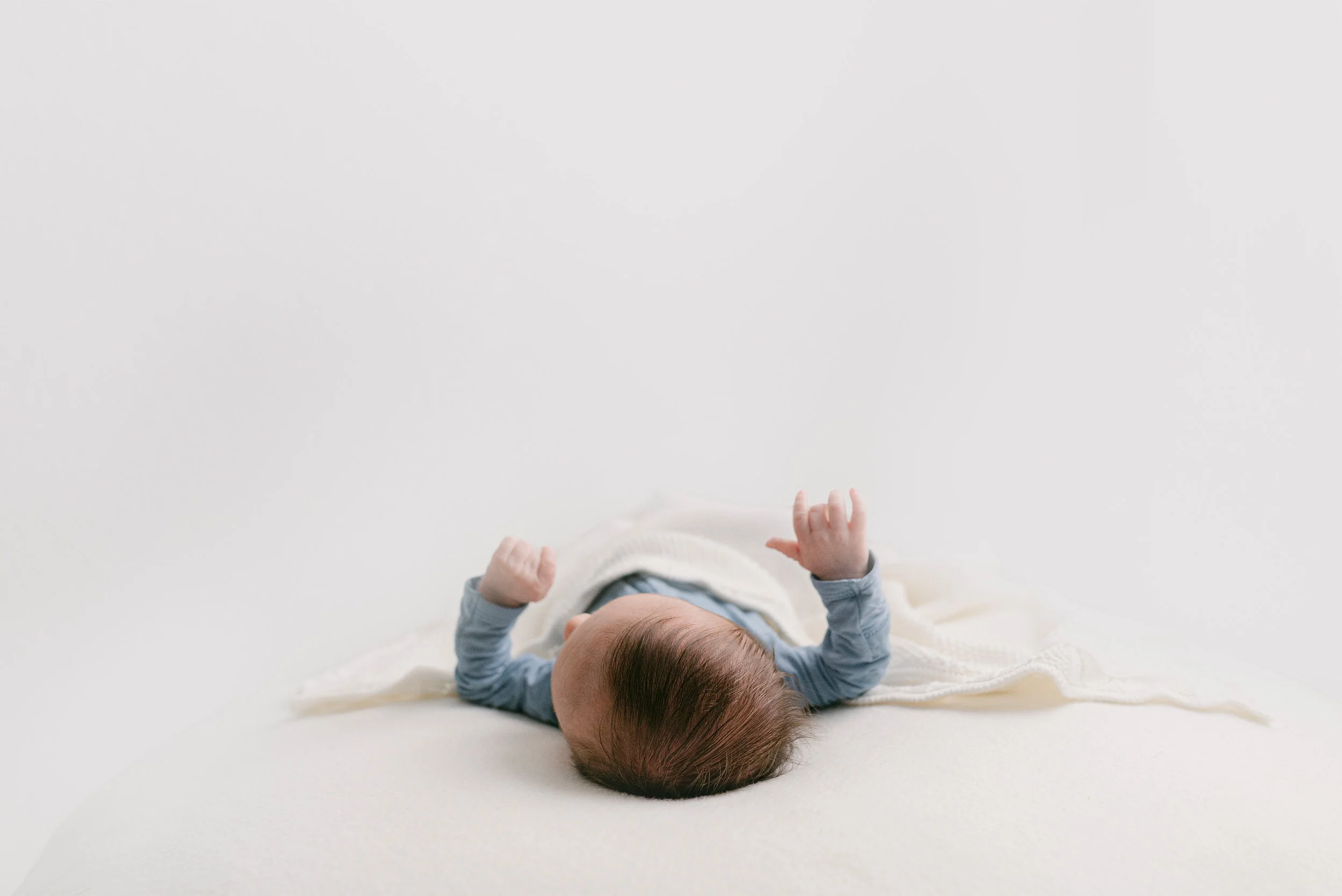 Newborn baby lying on back with arms raised under white blanket, award-winning baby photographer in Orange County