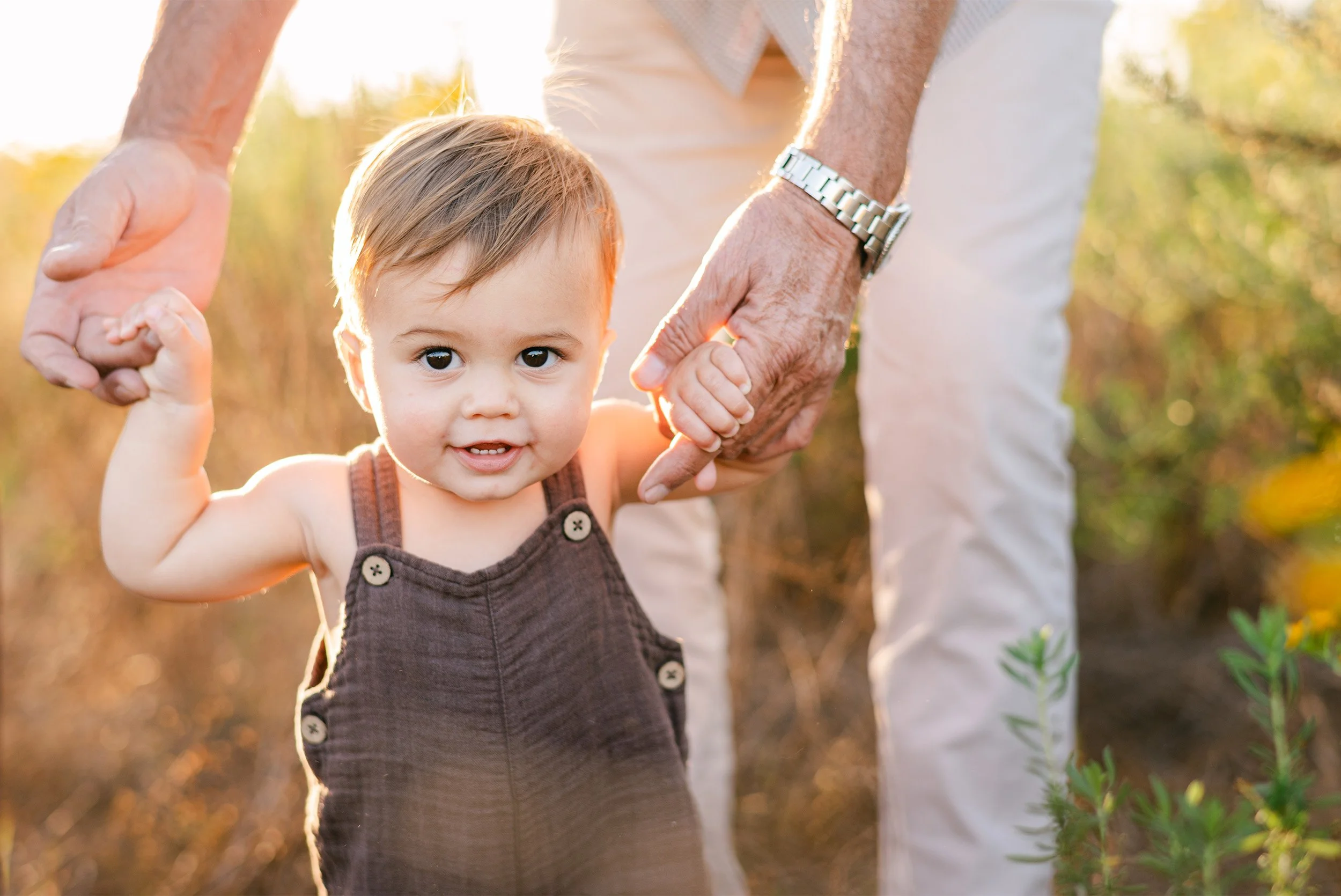 Award-winning family photographer in Orange County captures toddler holding grandfather's hand in sunlit field