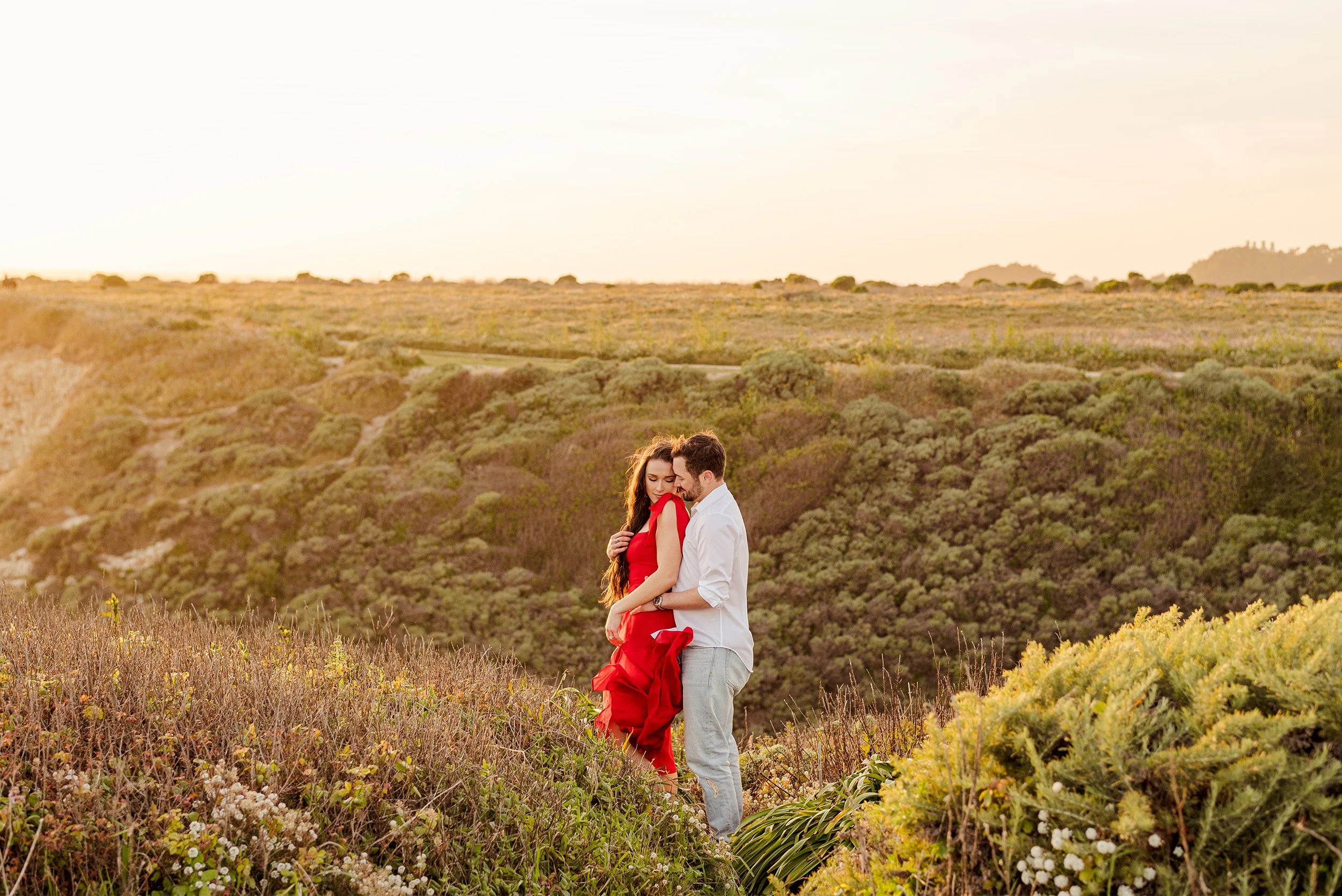 Couple with bride-to-be in red dress embracing on coastal bluff overlook in Santa Cruz, award-winning Santa Cruz engagement photographer