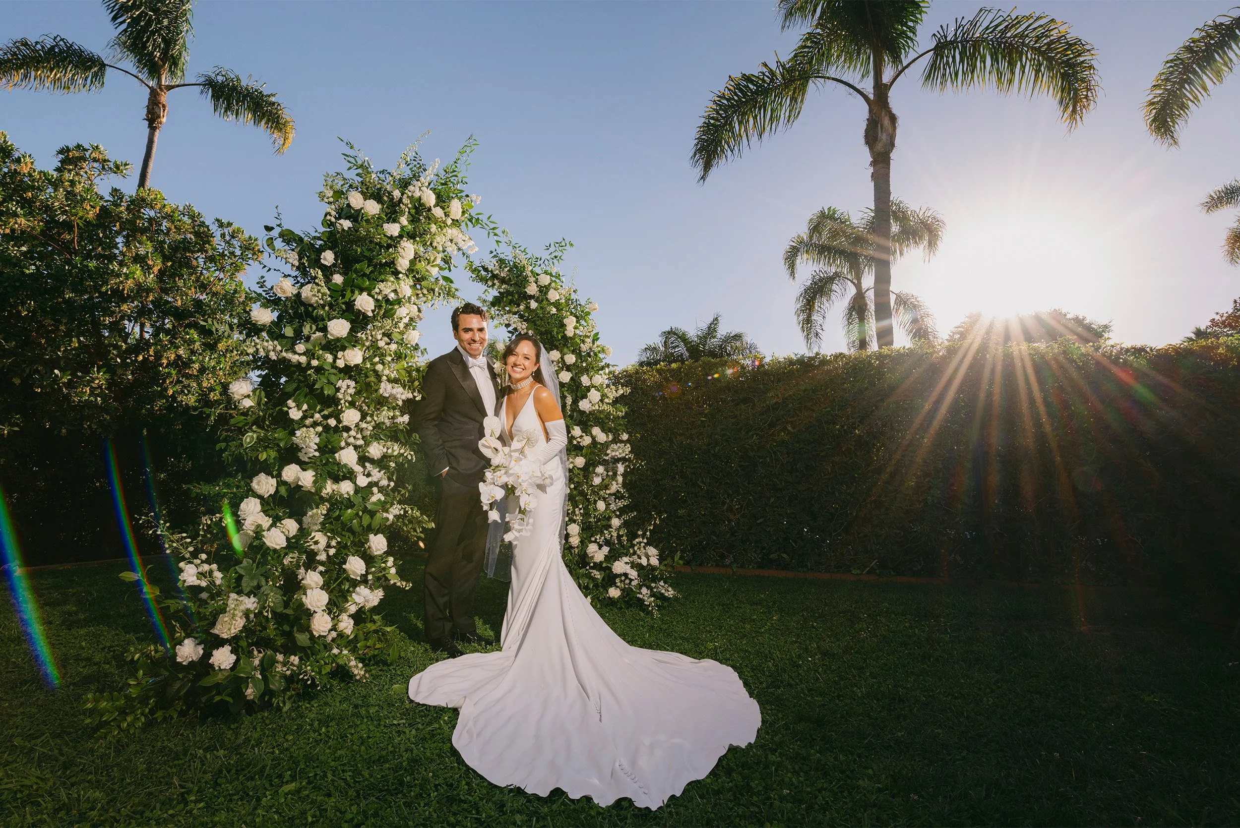 Orange County wedding photographer captures bride and groom golden hour in Santa Barbara portrait beneath white floral arch with palm trees