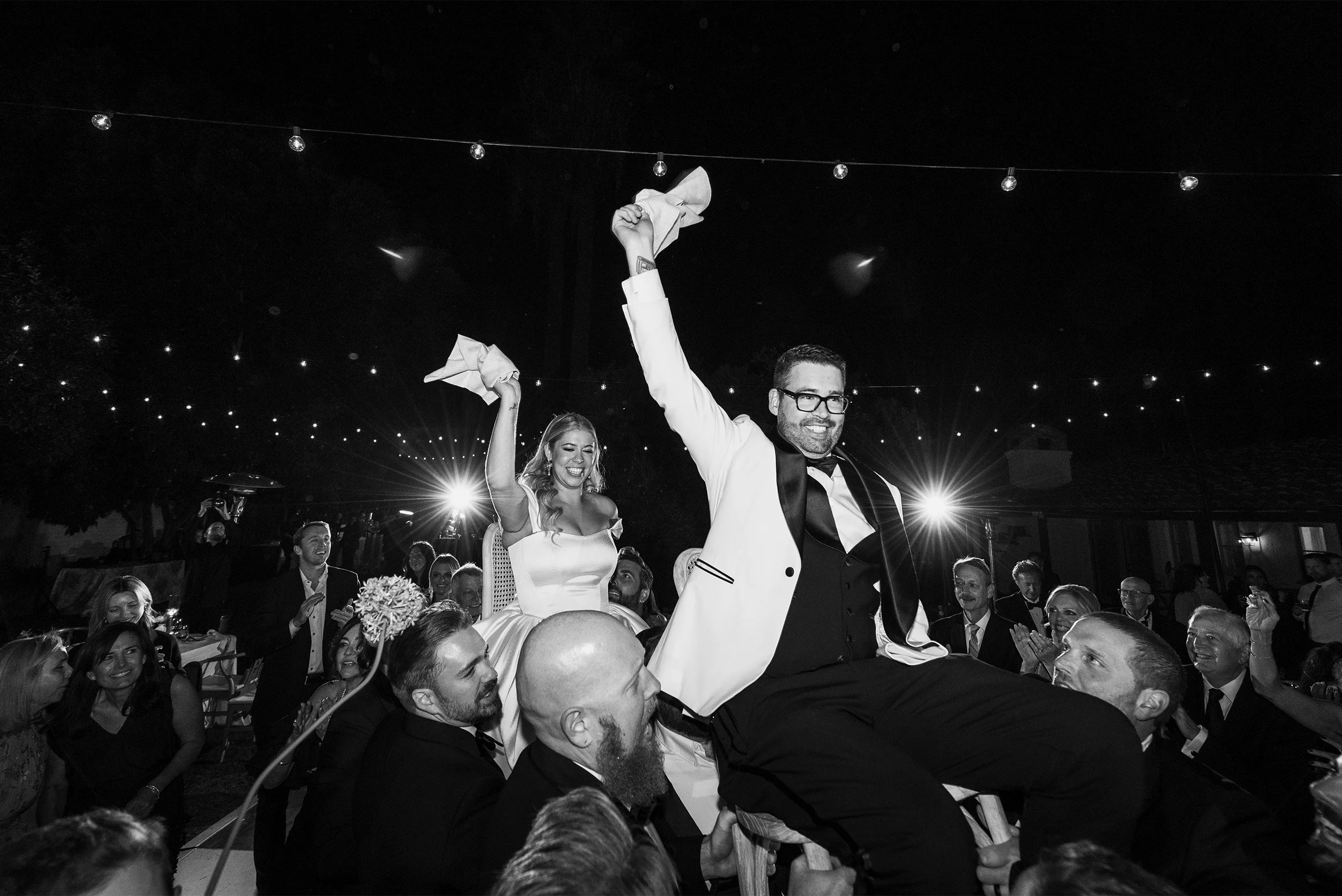 Wedding guests lifting bride and groom on chairs during reception celebration, black and white photo