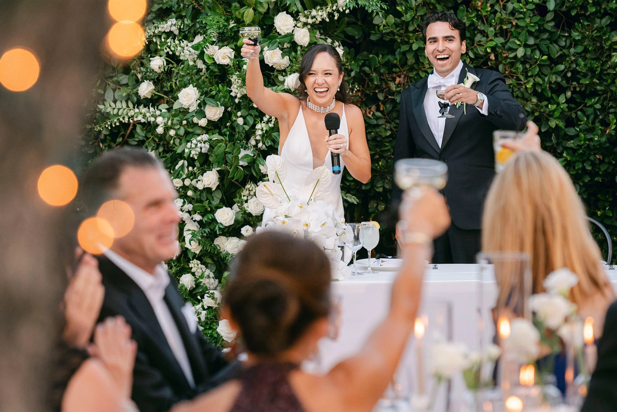 Highly recommended wedding photographer in Santa Barbara captures bride giving toast with microphone as groom raises glass among white rose backdrop