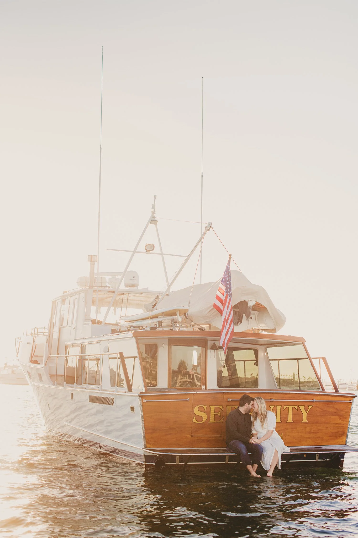 Couple sitting on the back of a vintage yacht named "Serenity" at sunset, Newport Beach harbor engagement session