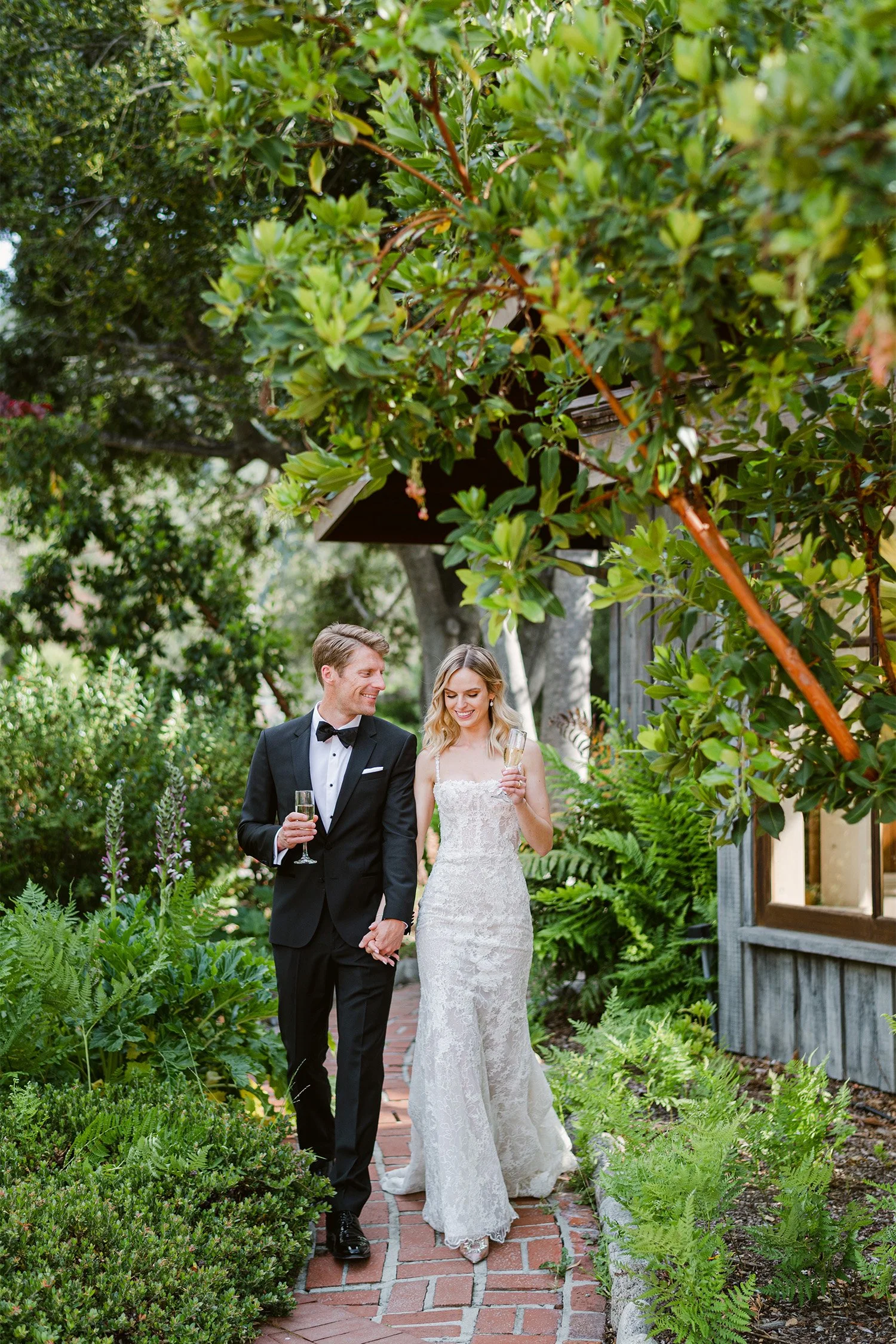 Orange County wedding photographer captures bride and groom walking through lush garden pathway with champagne toasts, Gardener Ranch wedding venue in Carmel Valley