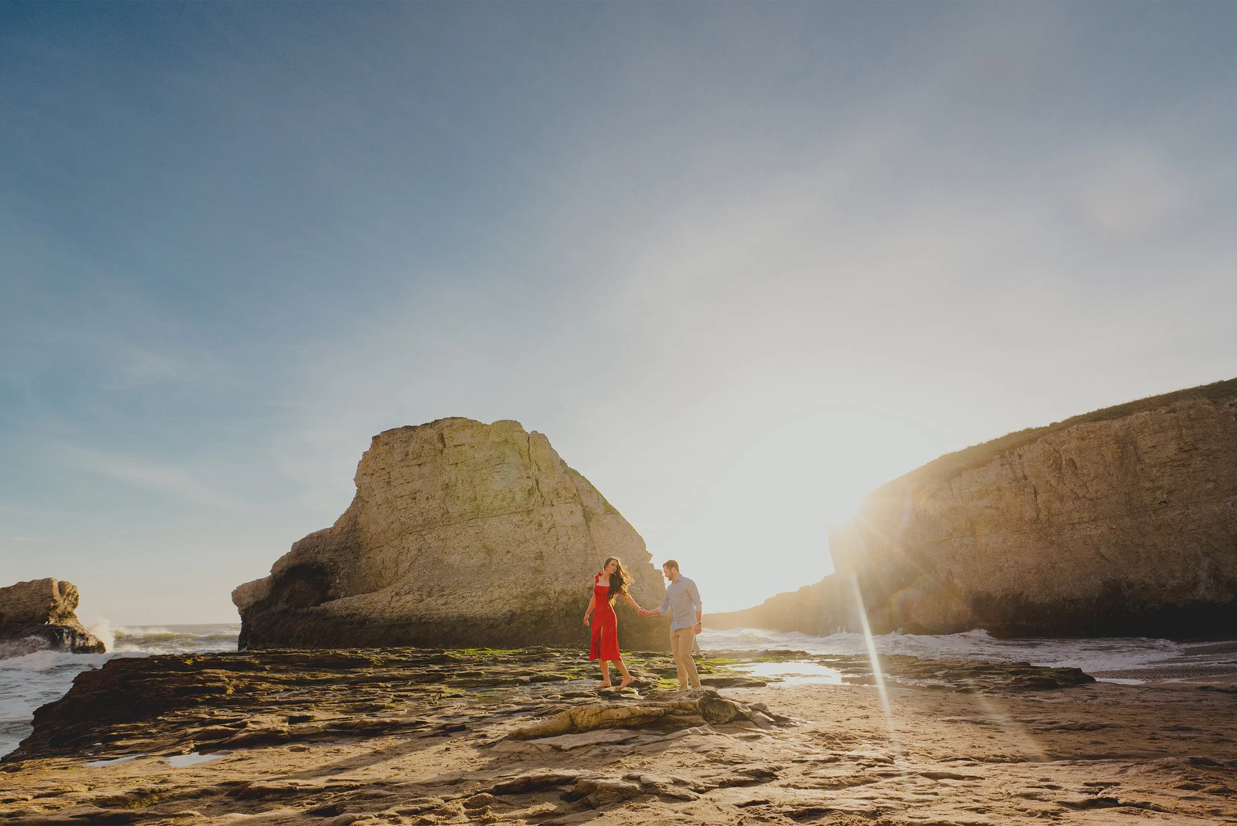 Couple with bride-to-be in red dress walking hand in hand past sea cliffs at sunset, highly recommended engagement photographer Orange County