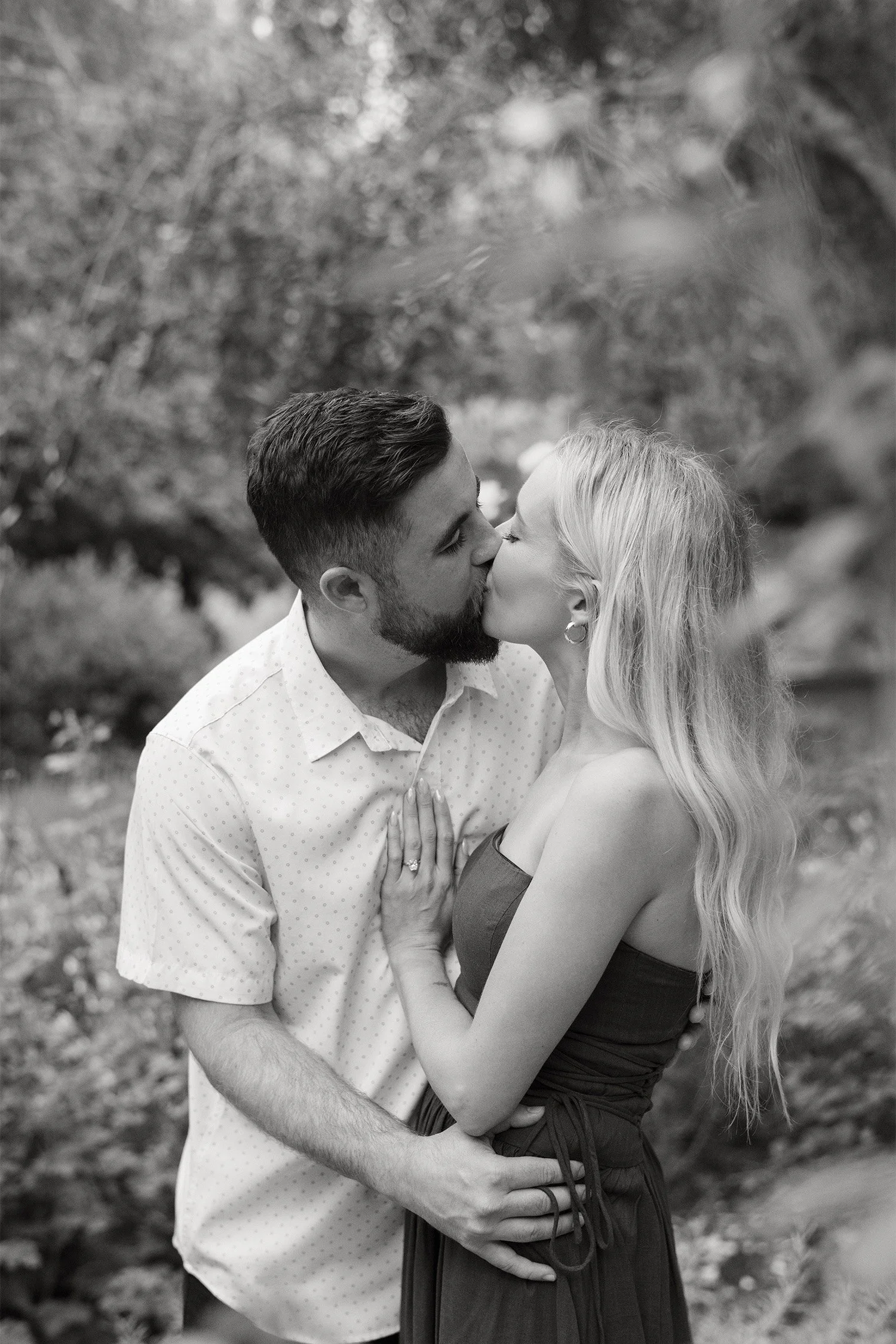 Orange County engagement photographer captures couple sharing a kiss under dappled tree light in black and white