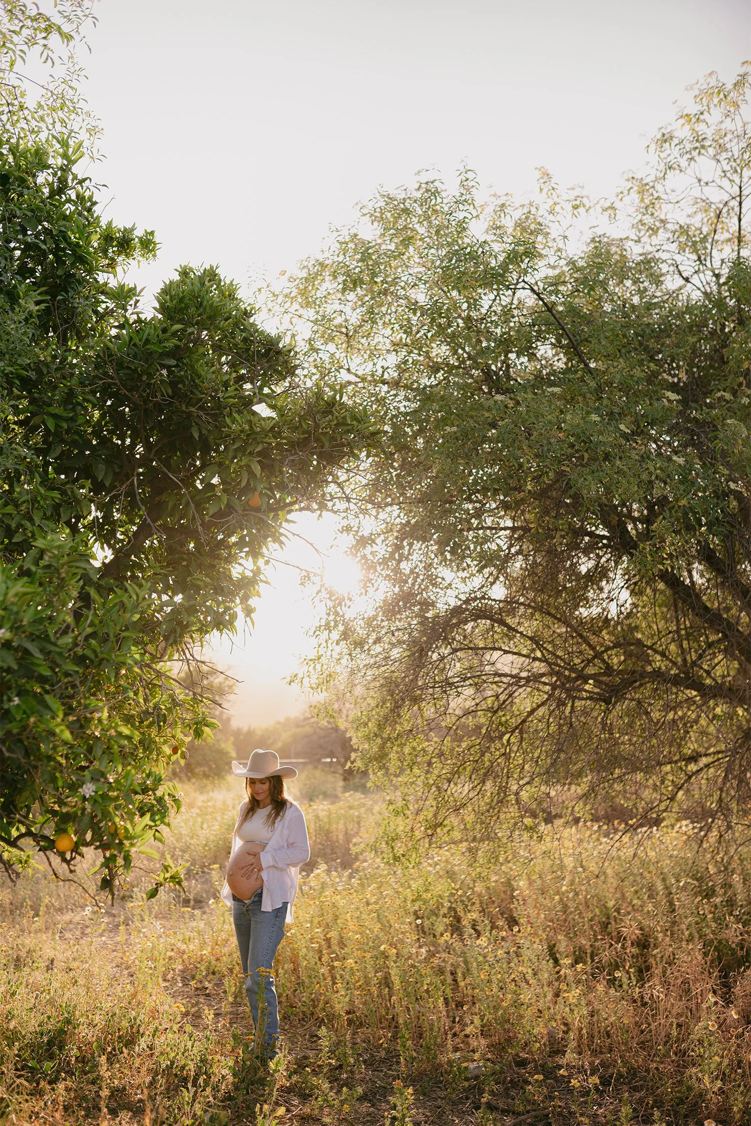 Pregnant woman in cowboy hat cradling belly in orchard at golden hour, Orange County maternity photographer