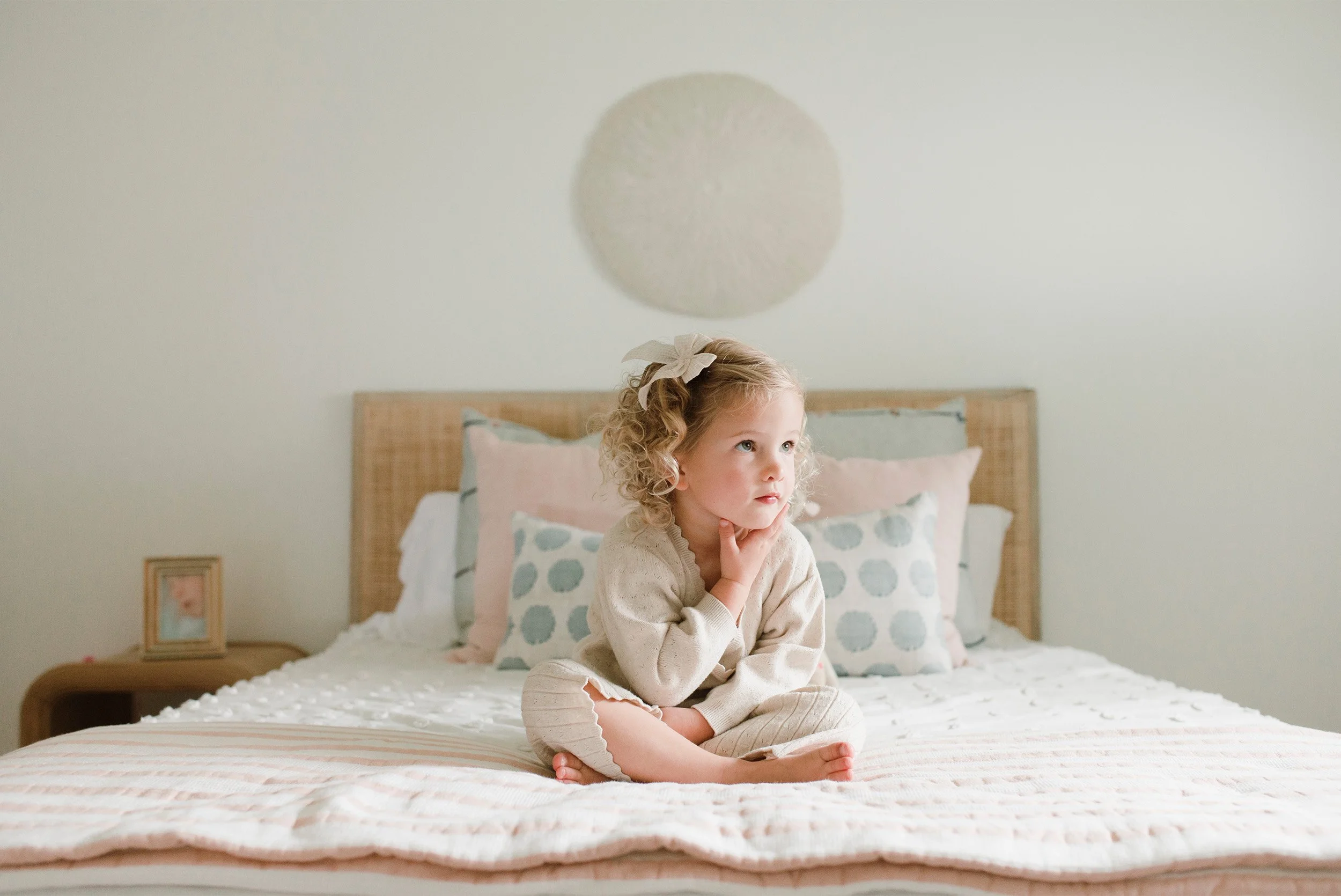 Award-winning family photographer in Orange County captures curly-haired girl sitting thoughtfully on bed at home