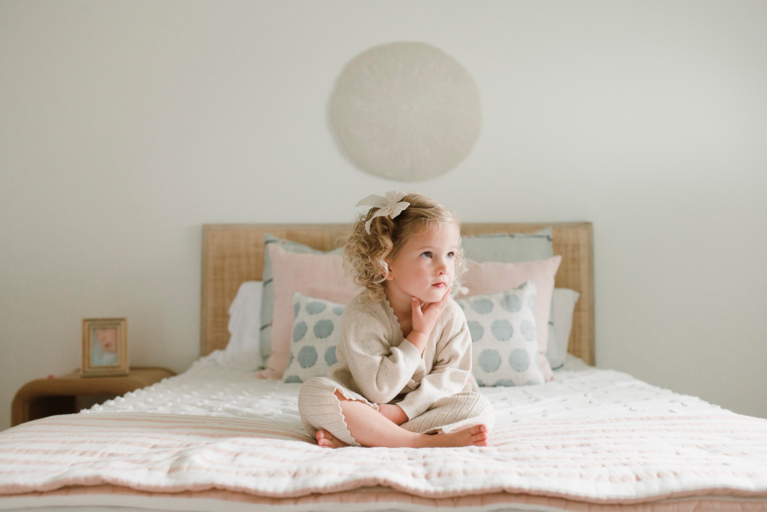 Award-winning family photographer in Orange County captures curly-haired girl sitting thoughtfully on her bed, in home family photos