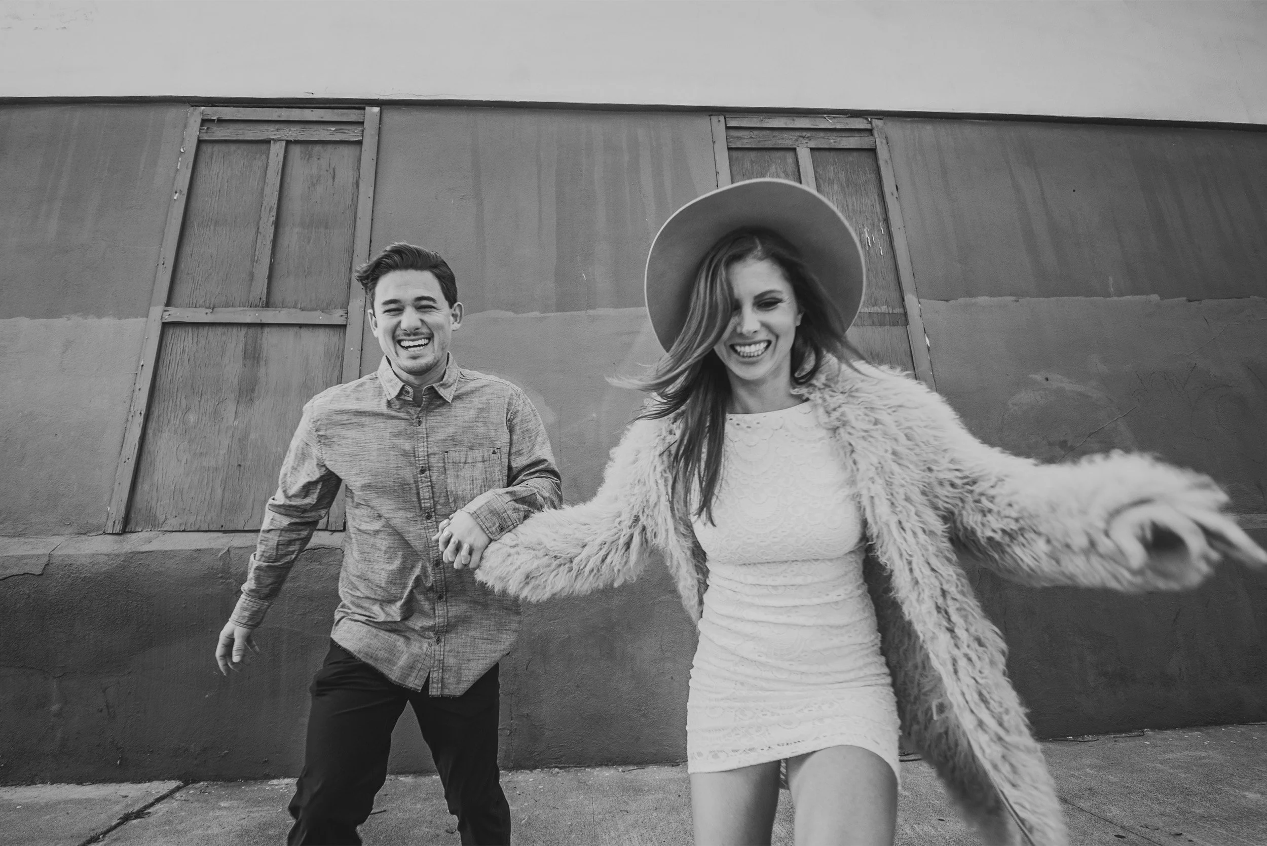 Black and white photo of laughing couple holding hands walking down an alley, candid Los Angeles engagement photography