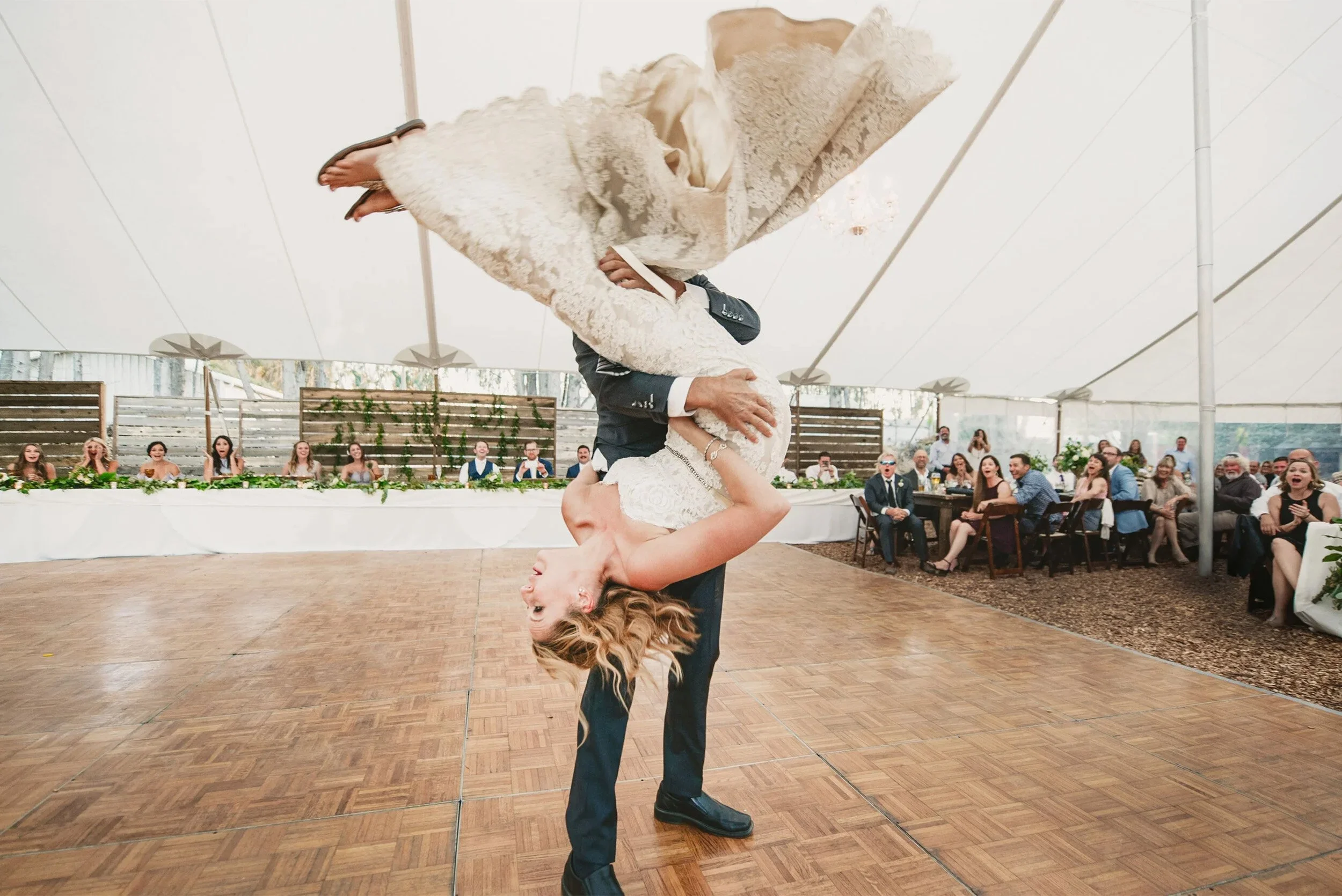 Groom flipping bride mid-dance under reception tent, guests cheering in background