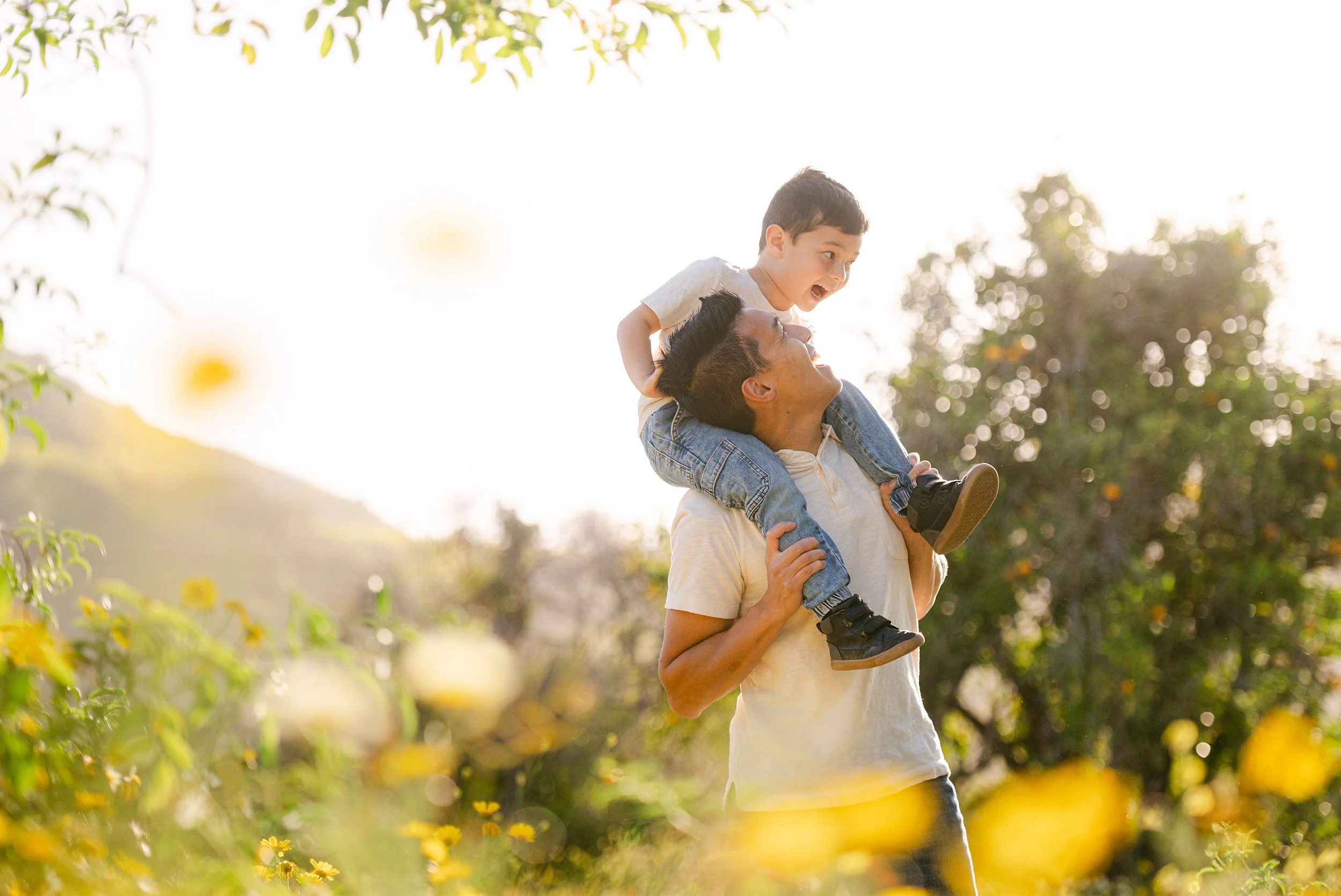 Award-winning family photographer in Orange County captures father lifting son playfully in sunlit flower field