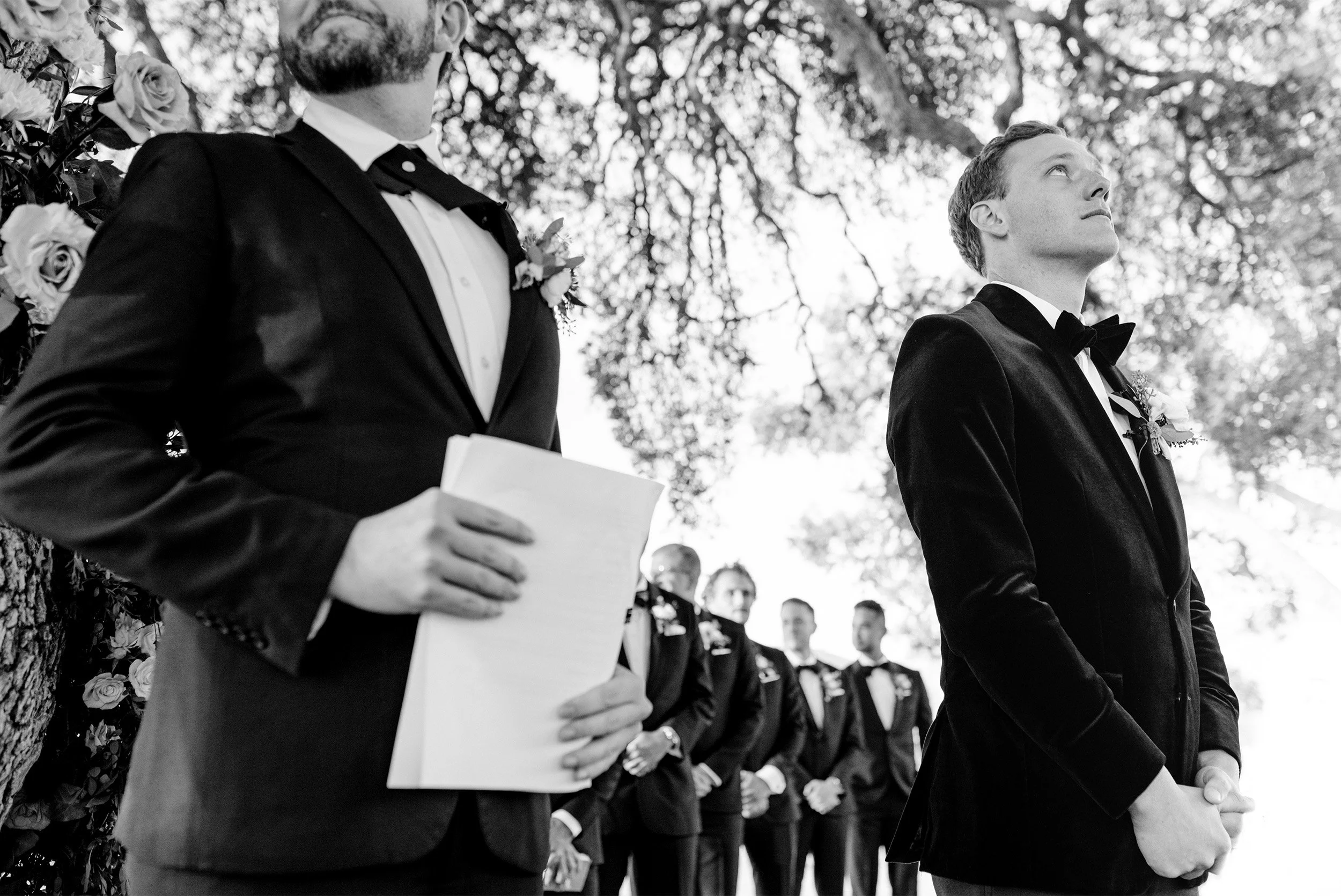 Groom and groomsmen in black tuxedos standing under oak tree during ceremony, black and white wedding photo