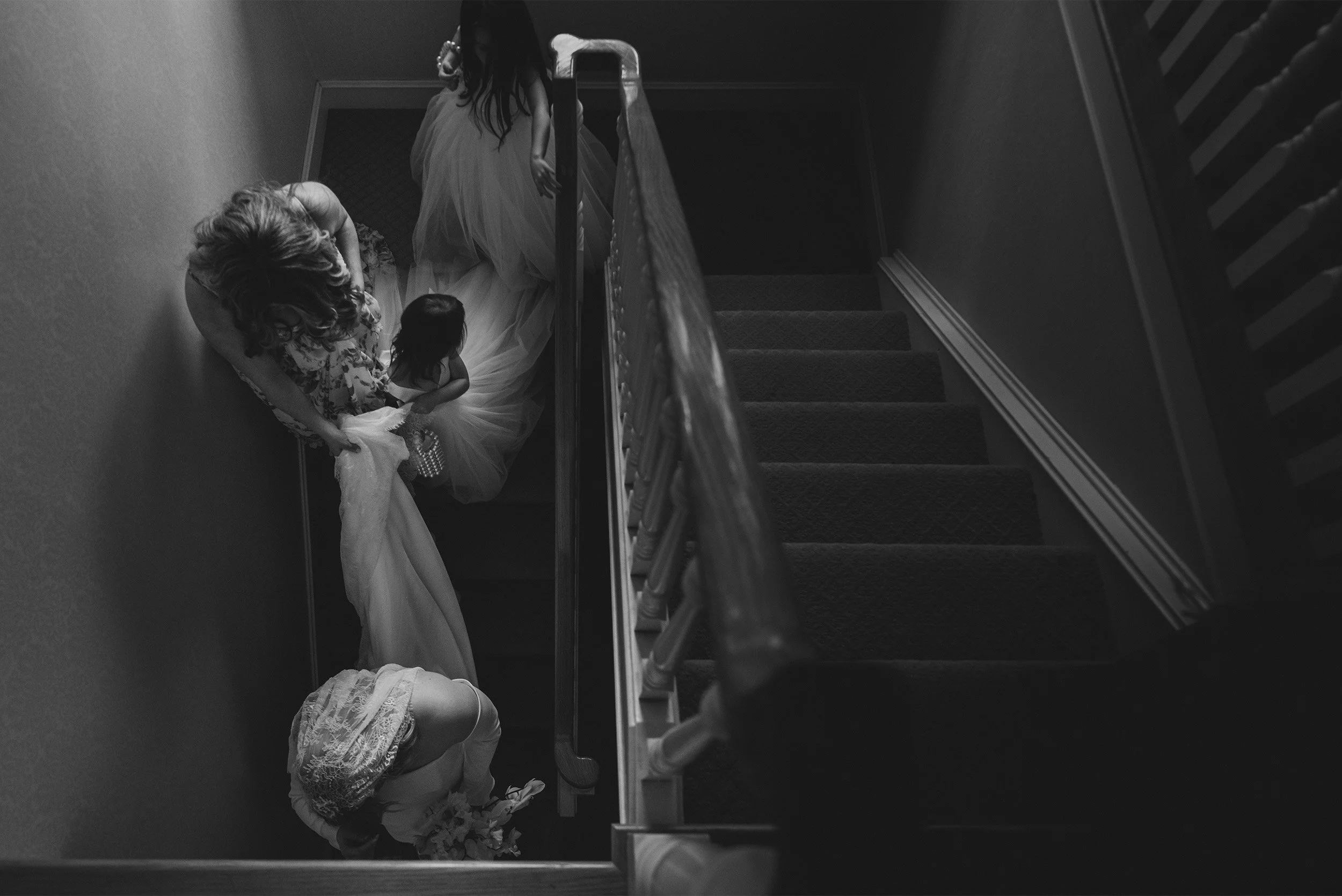 Highly recommended Orange County wedding photographer captures overhead view of bridesmaids helping bride down staircase with flower girl, black and white