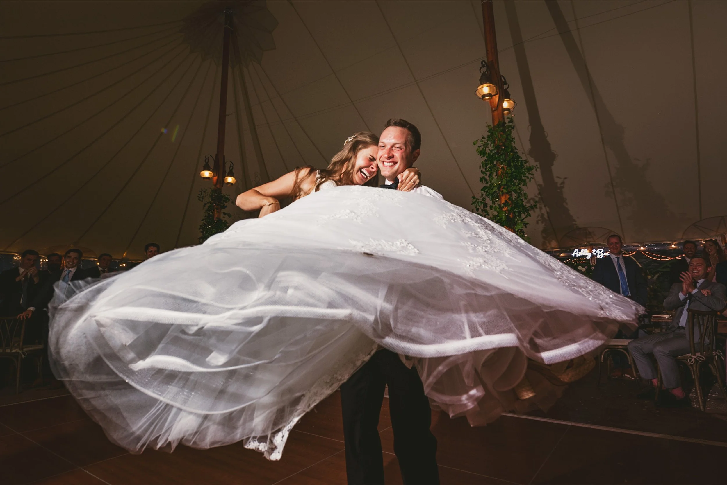 Groom lifting laughing bride as her wedding dress swirls under reception tent, Orange County wedding photography