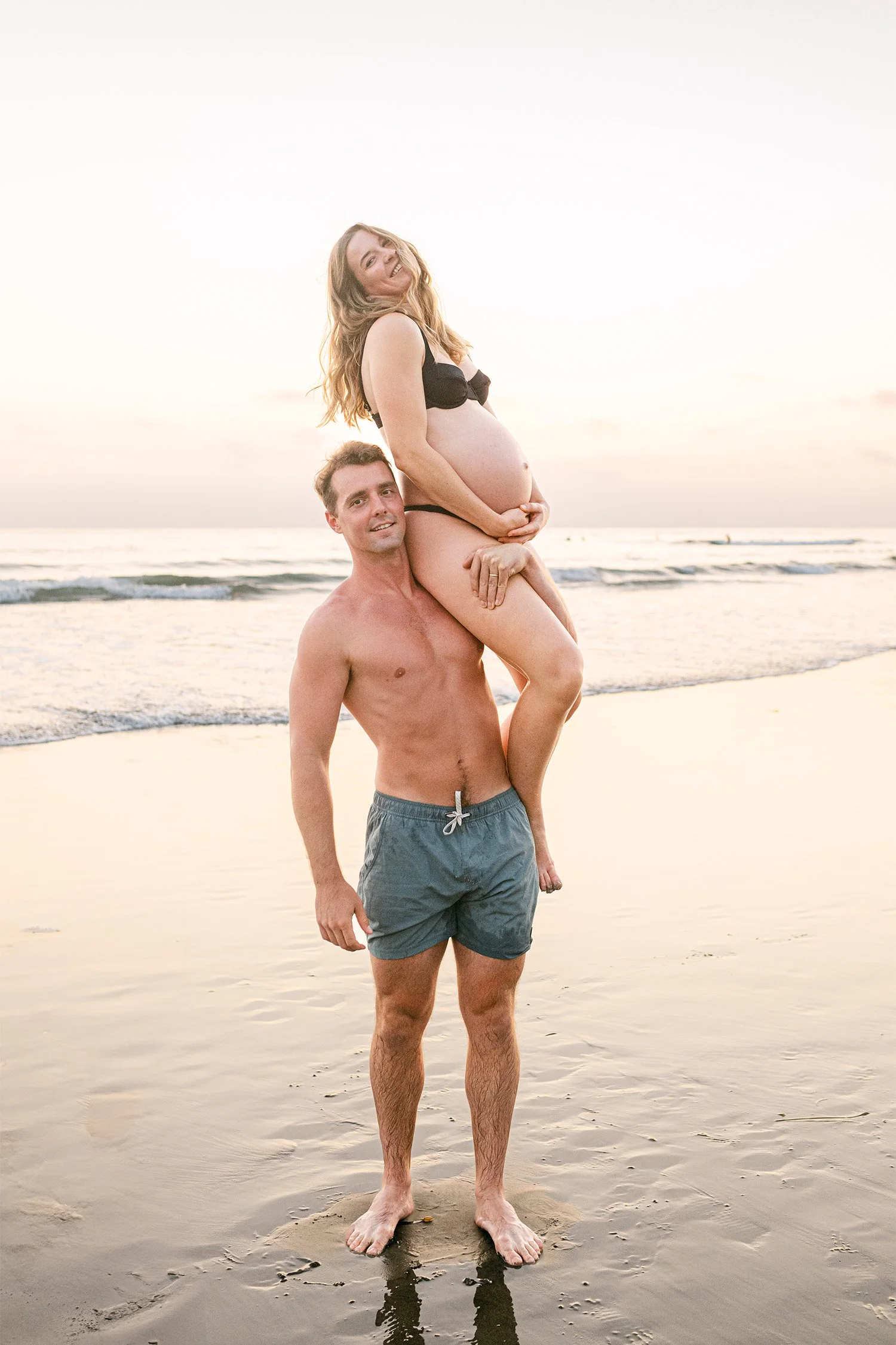 Pregnant woman laughing on partner's shoulders at sunset beach, award-winning maternity photographer in Orange County