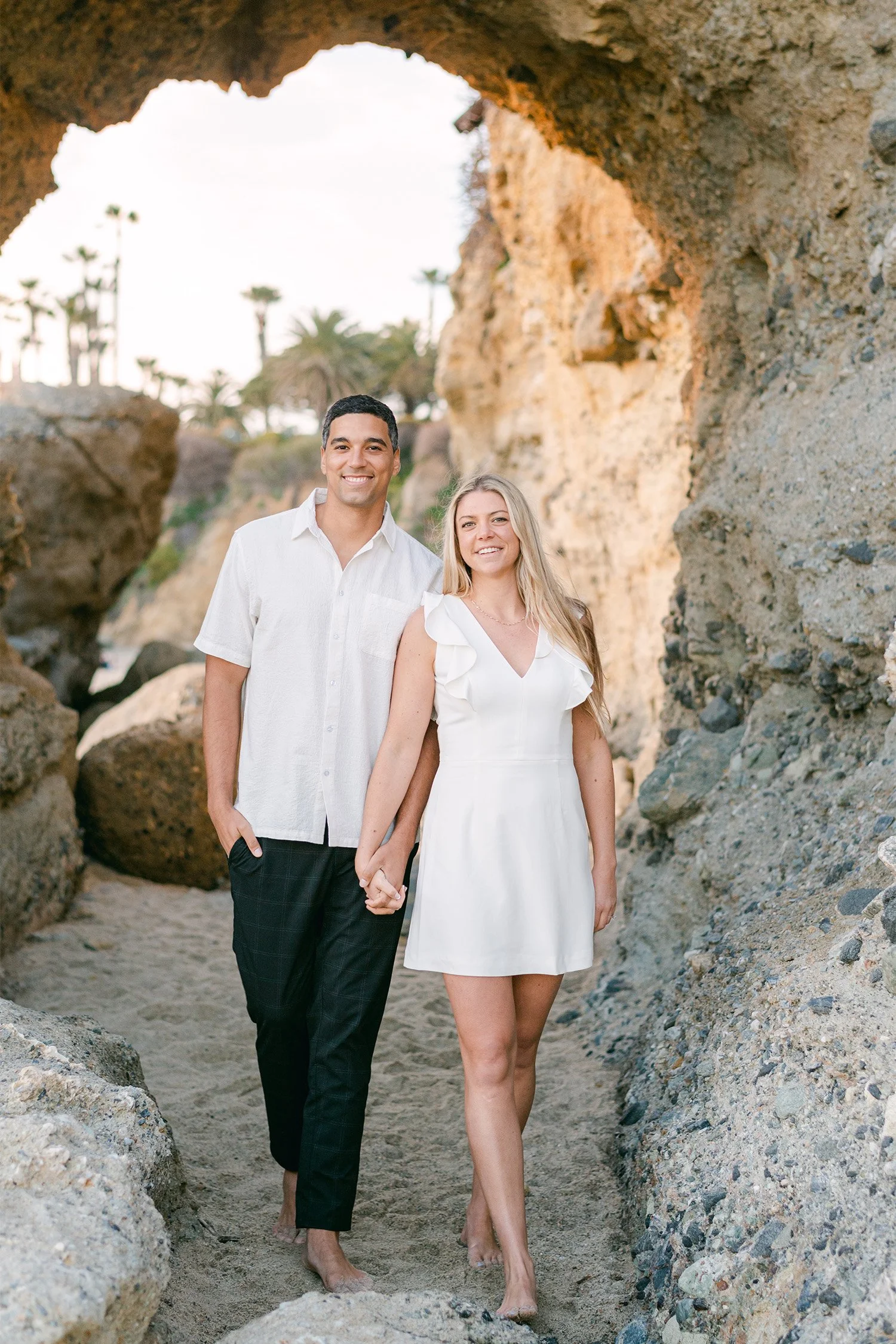Orange County engagement photographer captures couple holding hands beneath rock archway at Laguna Beach