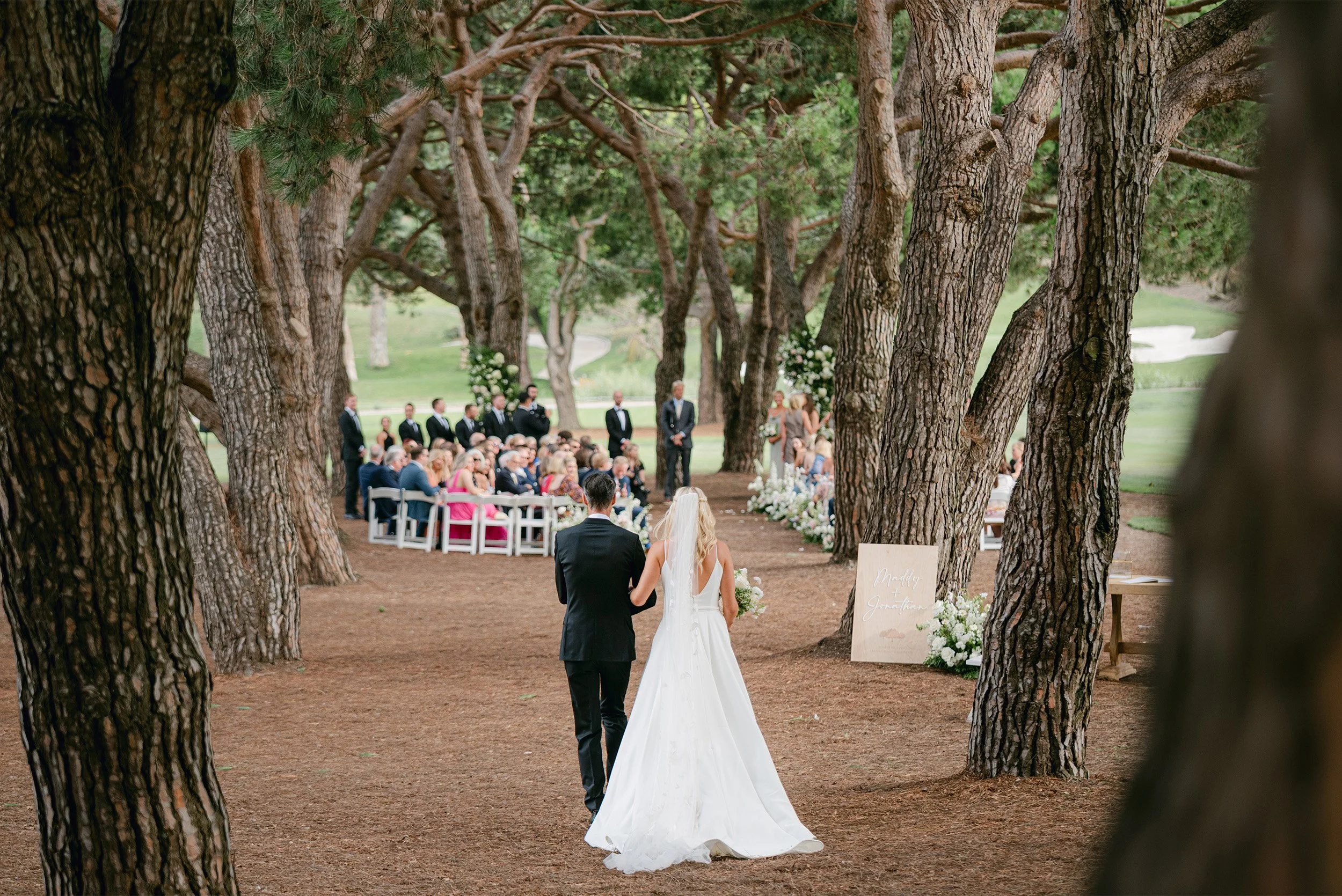 Bride and father of the bride walking down tree-lined aisle toward guests at The Ranch Laguna Beach, an outdoor wedding ceremony