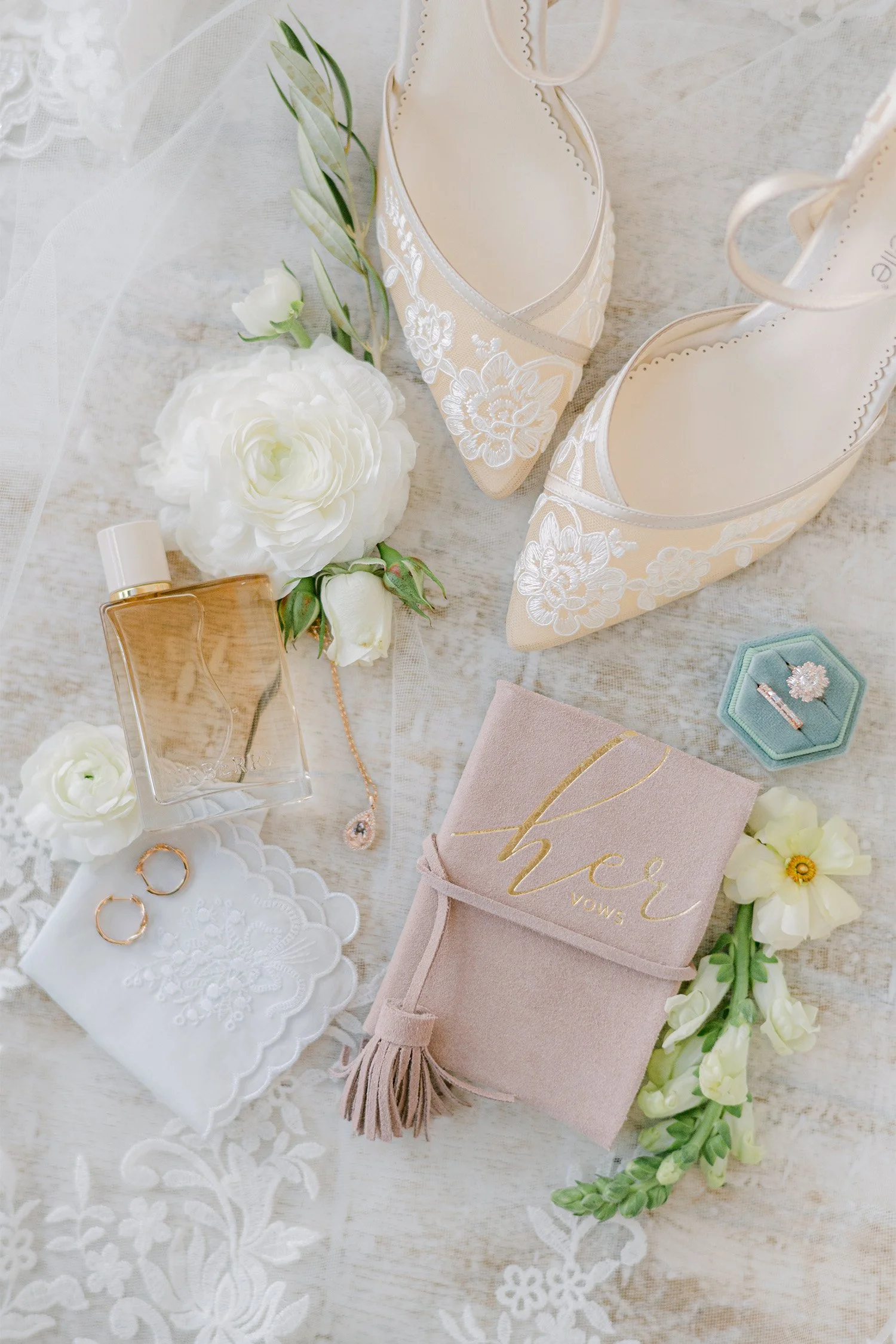 Bridal flat lay with lace wedding shoes, perfume, rings, and vow book, wedding detail photography