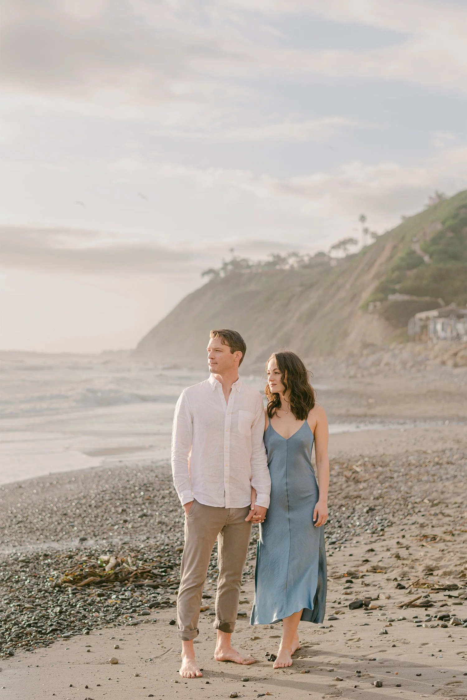 Orange County engagement photographer captures couple walking hand in hand along rocky beach beneath coastal bluffs