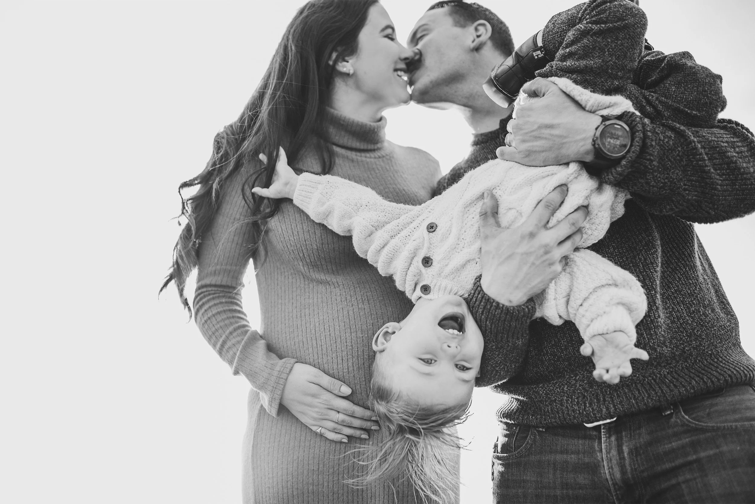 Award-winning Orange County family photographer captures playful black and white moment of parents kissing while holding their child