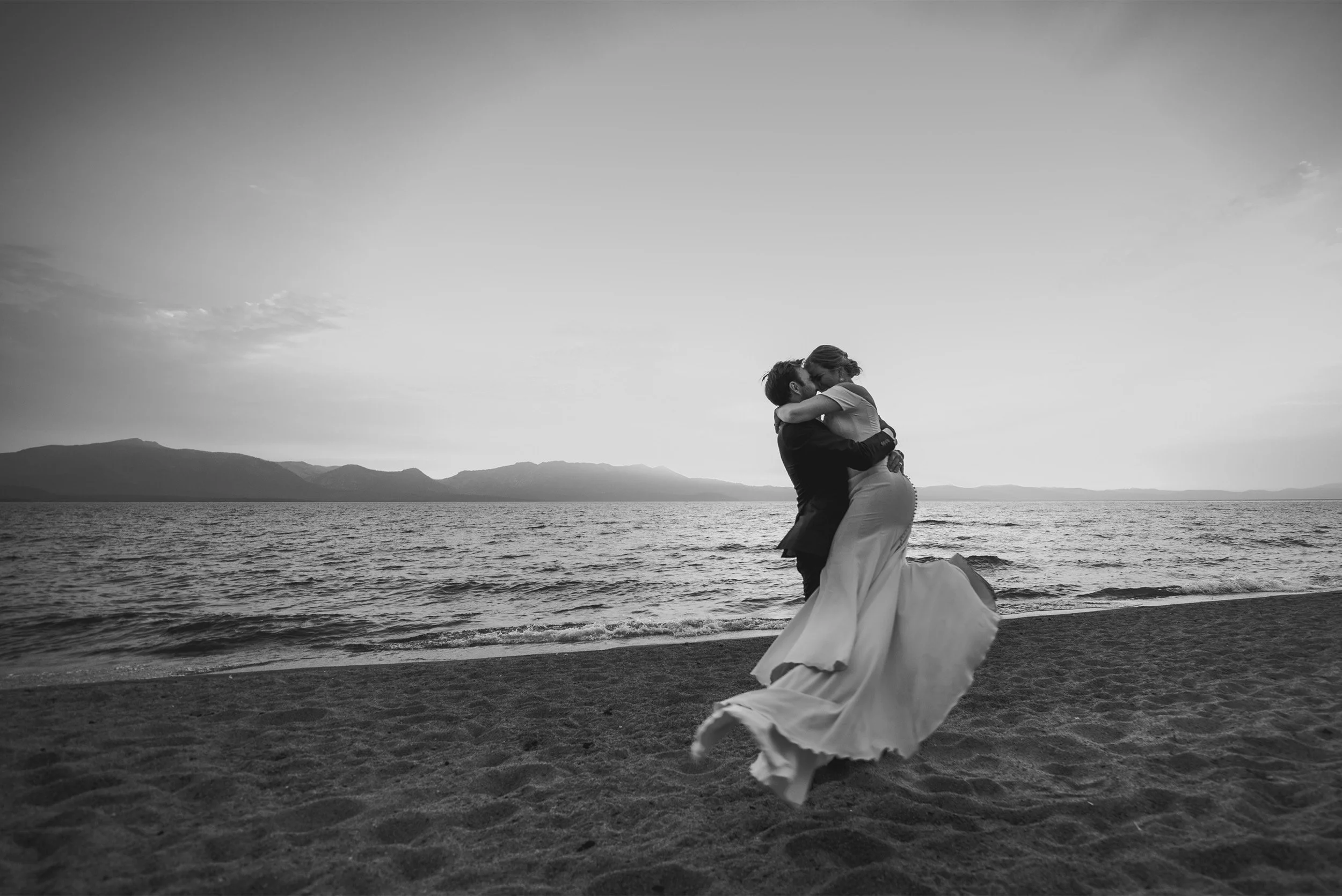 Groom lifting bride on the beach at sunset, black and white Lake Tahoe wedding photo