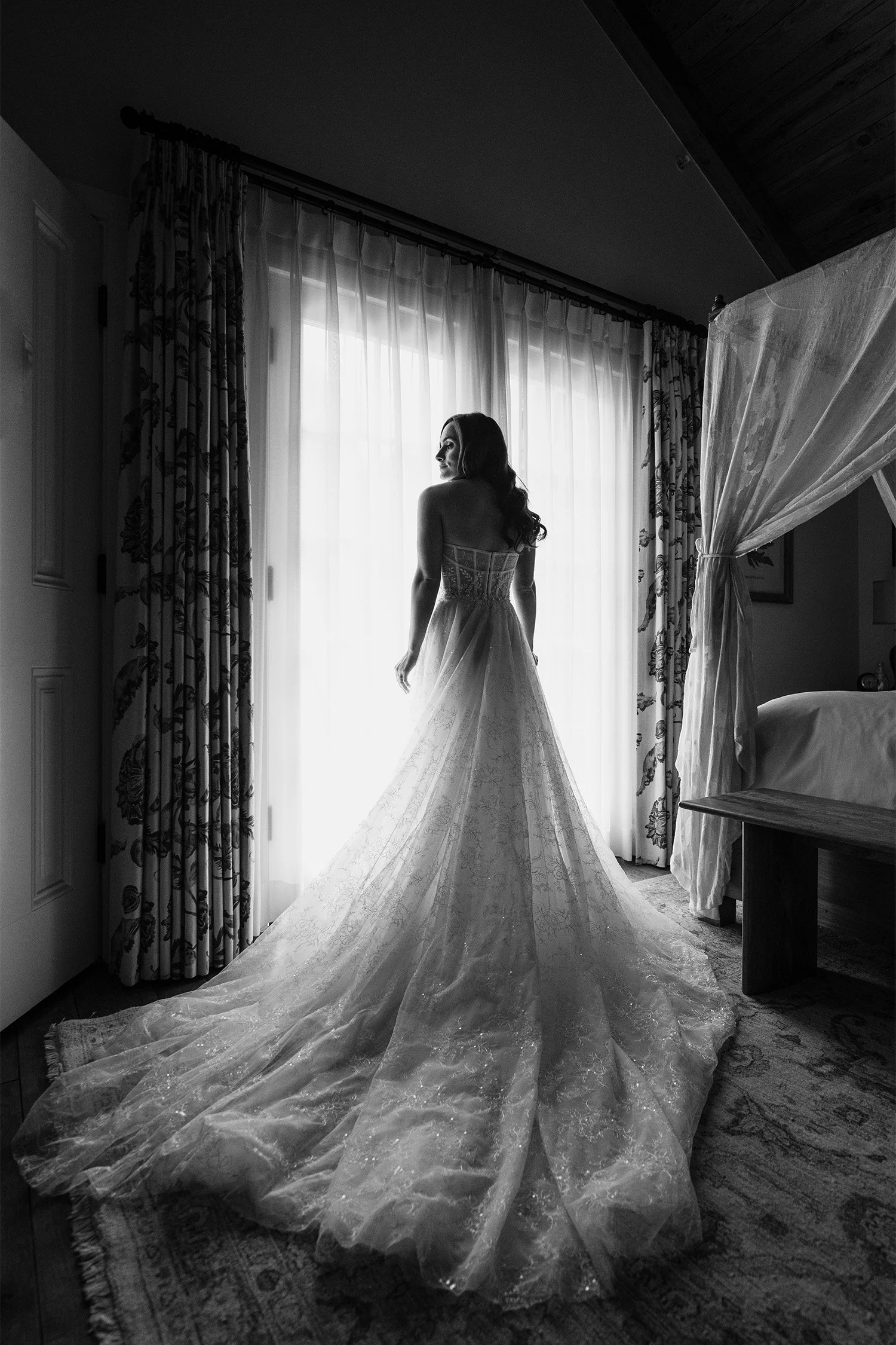 Bride in strapless lace gown silhouetted against sheer curtains, black and white bridal portrait