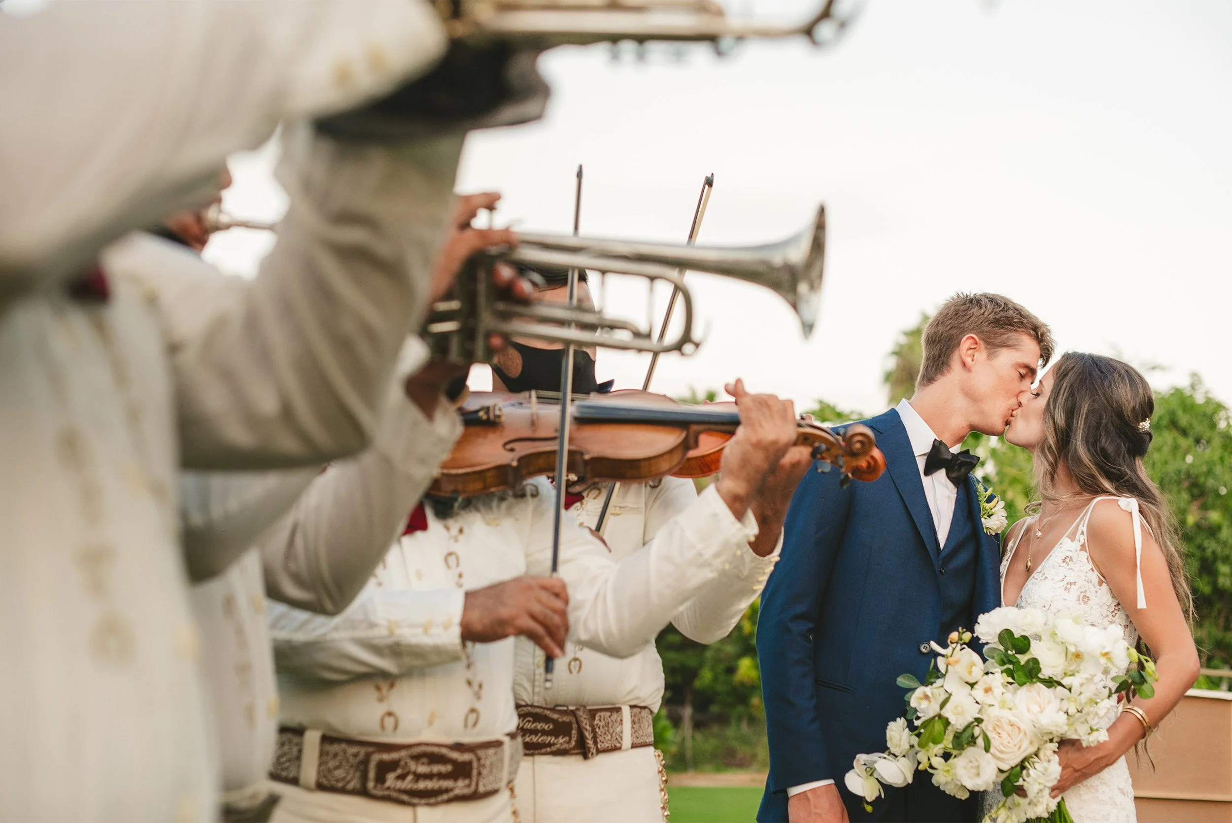 Newlyweds' first kiss framed by mariachi band musicians playing violin and trumpet at Flora Farms in Cabo, Mexico, International wedding photographer from Orange County