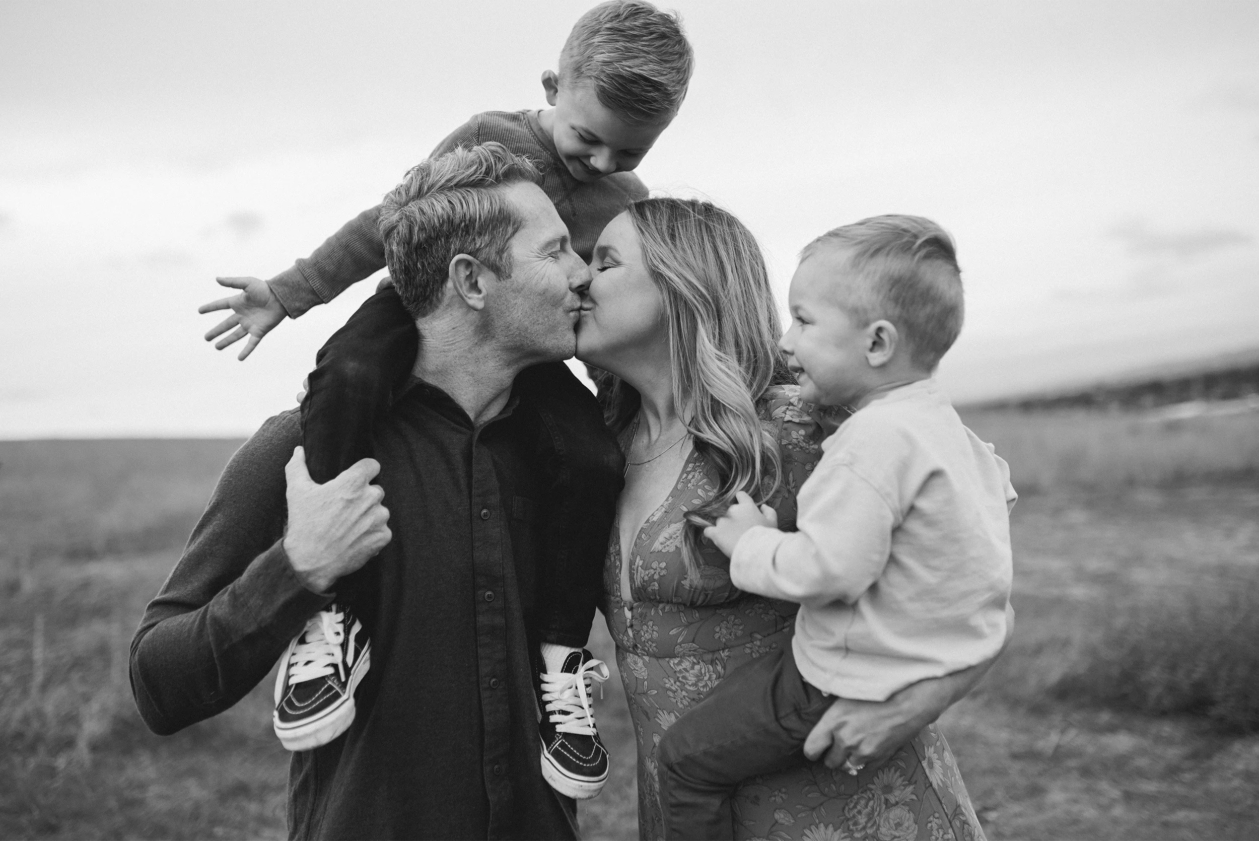 Candid moment of parents kissing while carrying two young sons in an open field, Orange County family photographer