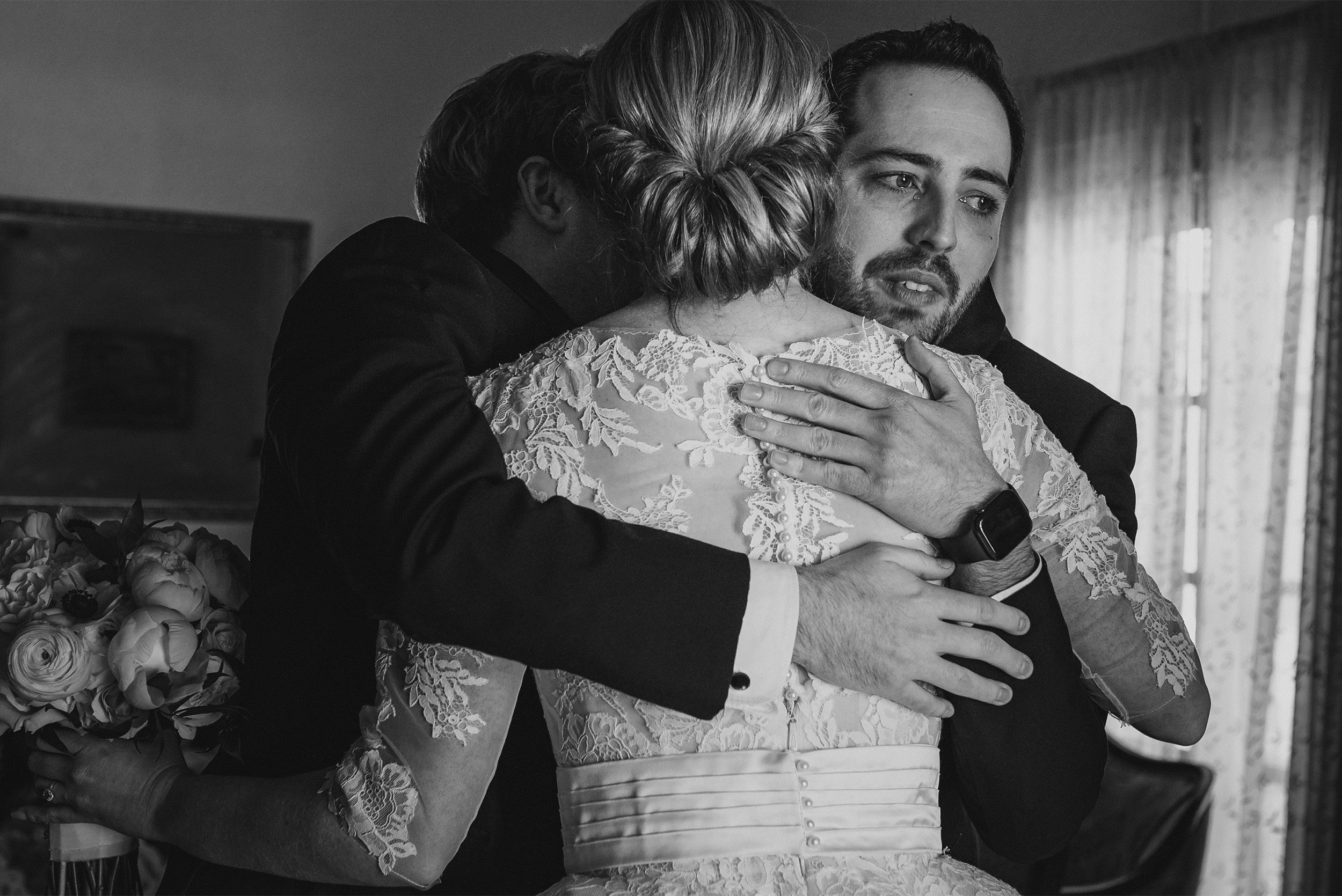 Bride and groom embracing family member during emotional moment, black and white candid wedding photo