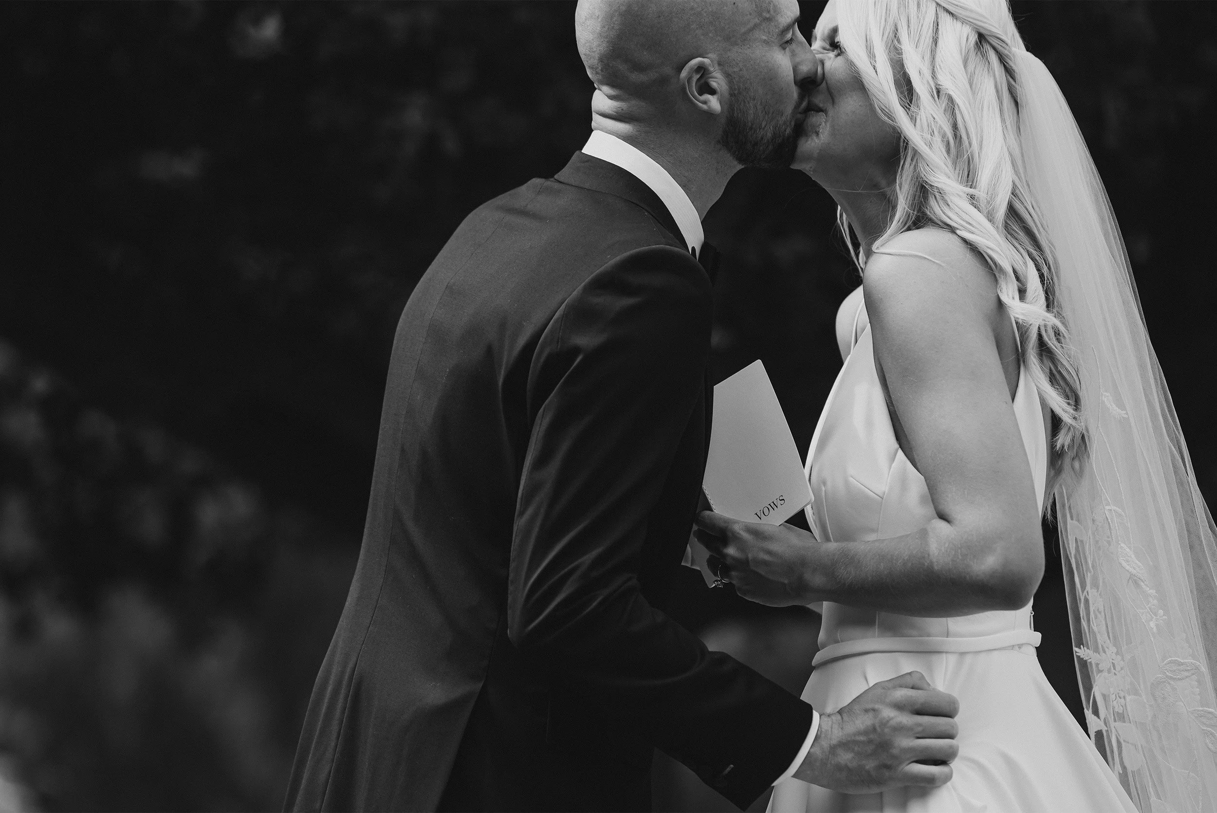 Award-winning Orange County wedding photographer captures intimate profile kiss between bride and groom holding vows cards at The Ranch at Laguna Beach, black and white