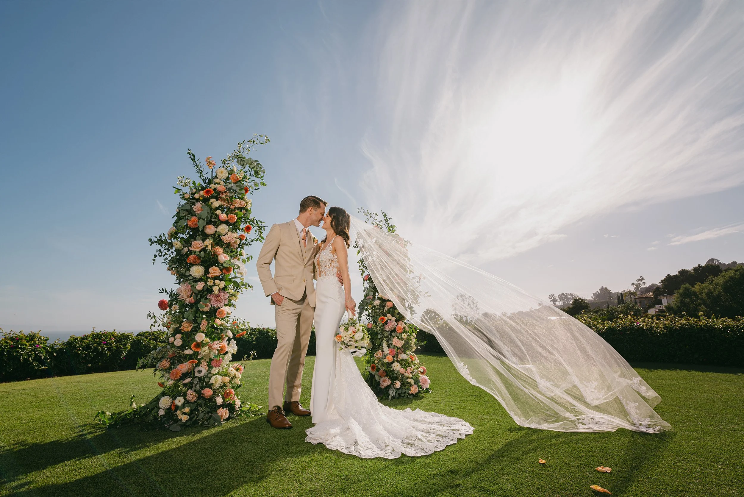 Santa Barbara wedding photographer captures bride and groom's ceremony kiss beneath dramatic sky with peach and white floral arch