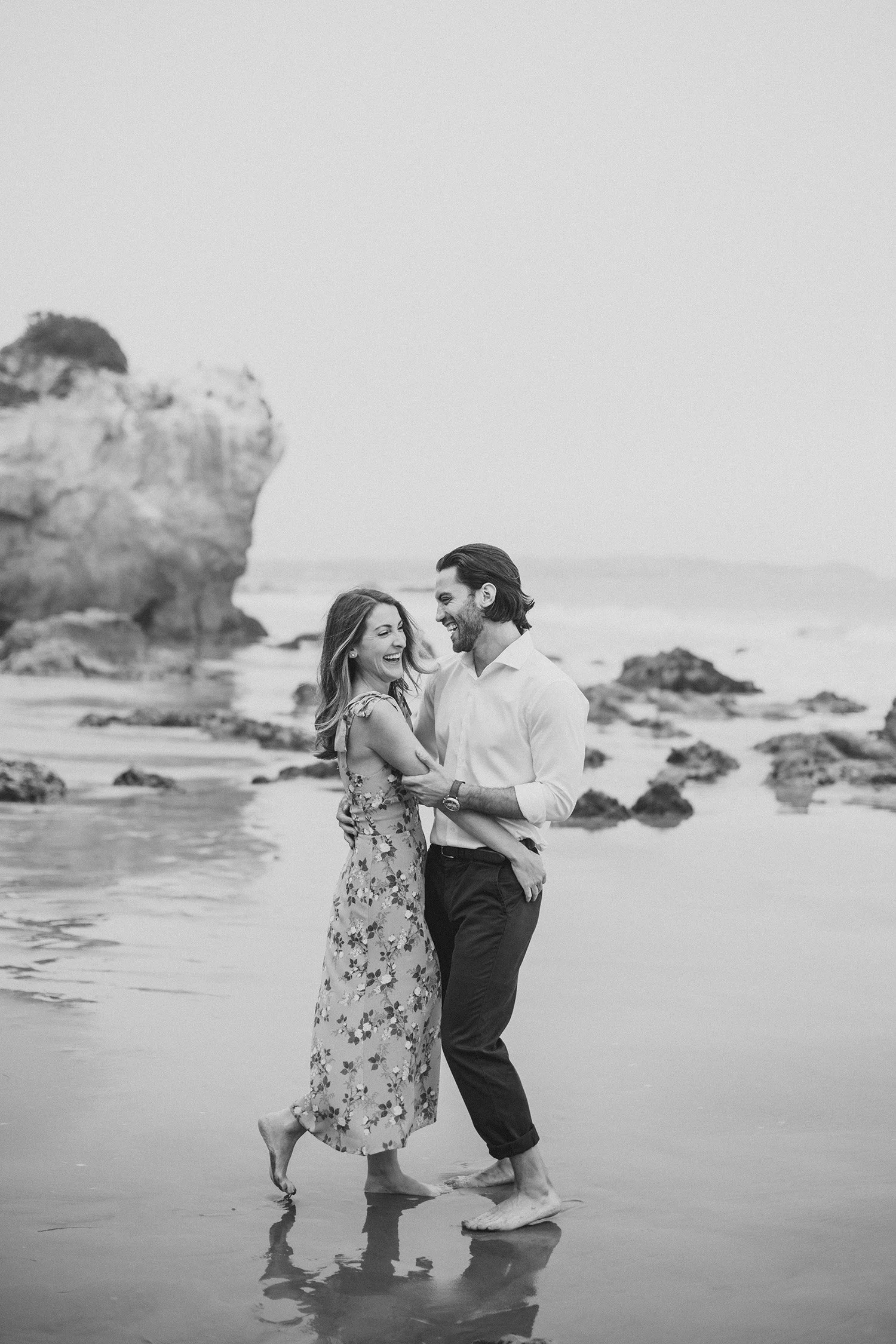 Black and white photo of laughing couple embracing barefoot on the beach near coastal rock formations, Malibu engagement photography