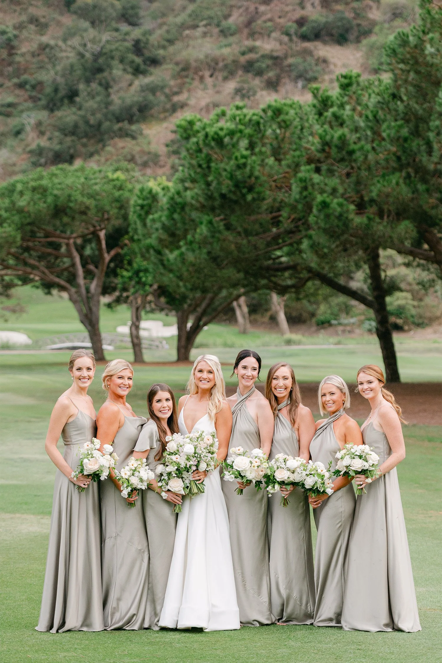 Bride and bridesmaids in sage green gowns posing together on golf course lawn at The Ranch Laguna Beach