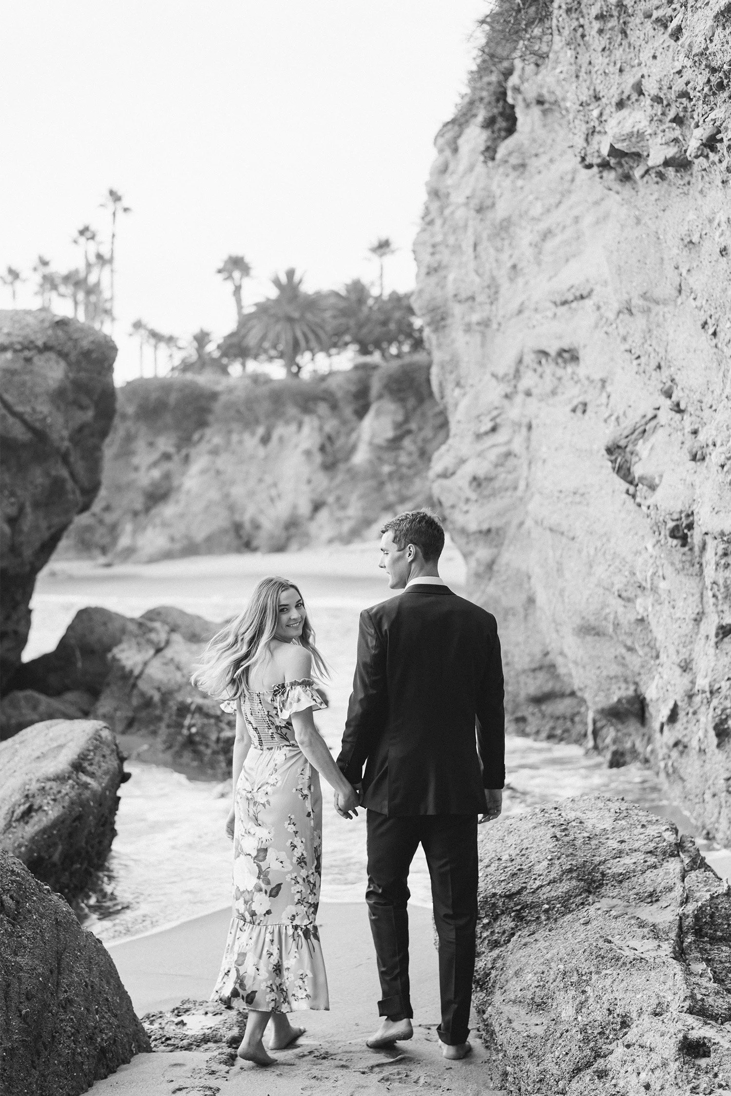 Orange County engagement photographer captures couple walking hand in hand through rocky beach cove in black and white