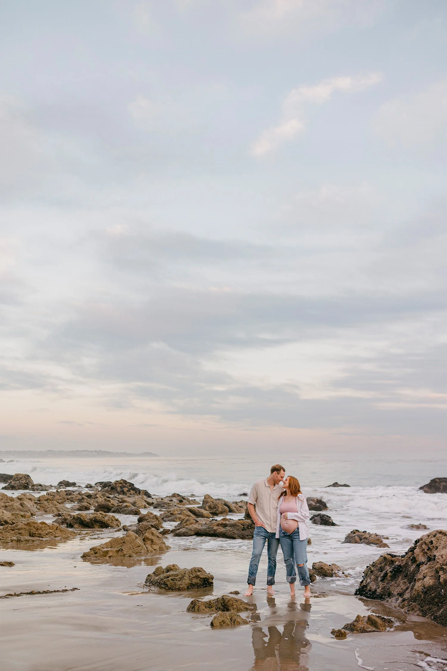 Pregnant couple embracing on rocky beach at sunset, Orange County maternity photographer