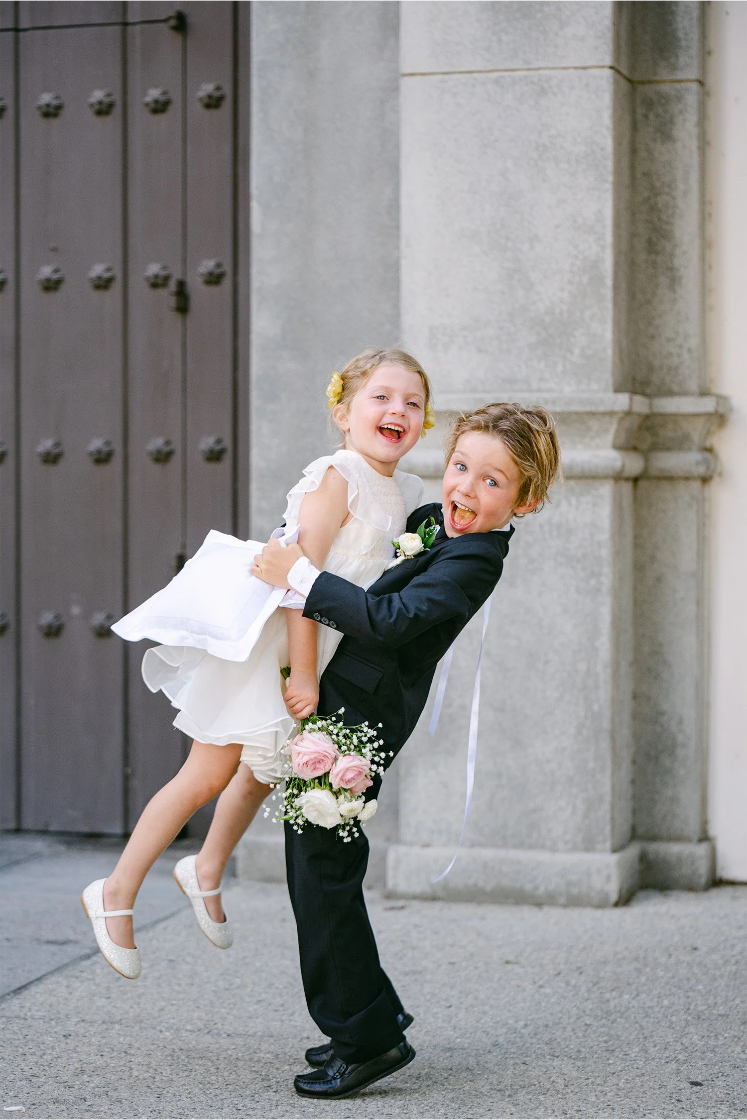 Ring bearer carrying flower girl piggyback style outside church doors, candid wedding photo