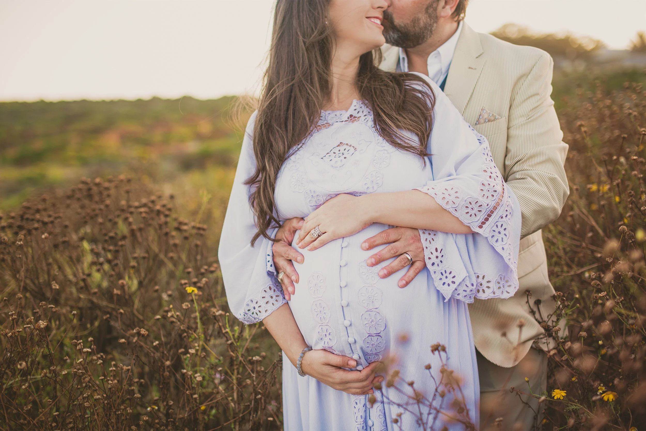 Orange County maternity photographer captures close-up of couple embracing pregnant belly in golden wildflower field