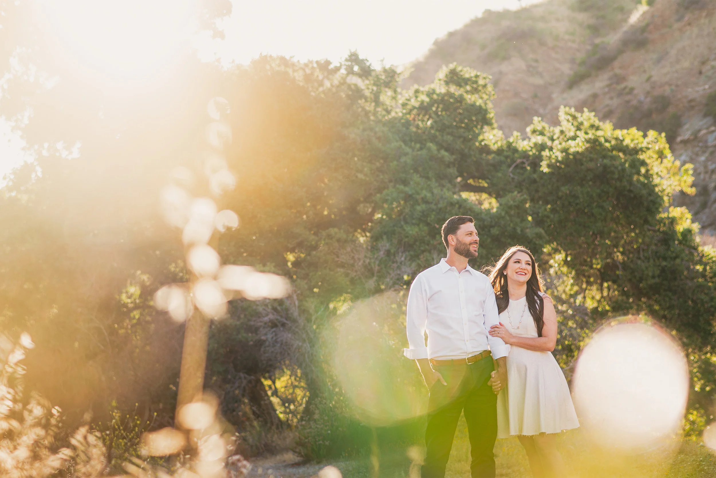 Couple smiling together with sun flare through trees, highly recommended engagement photographer in Orange County